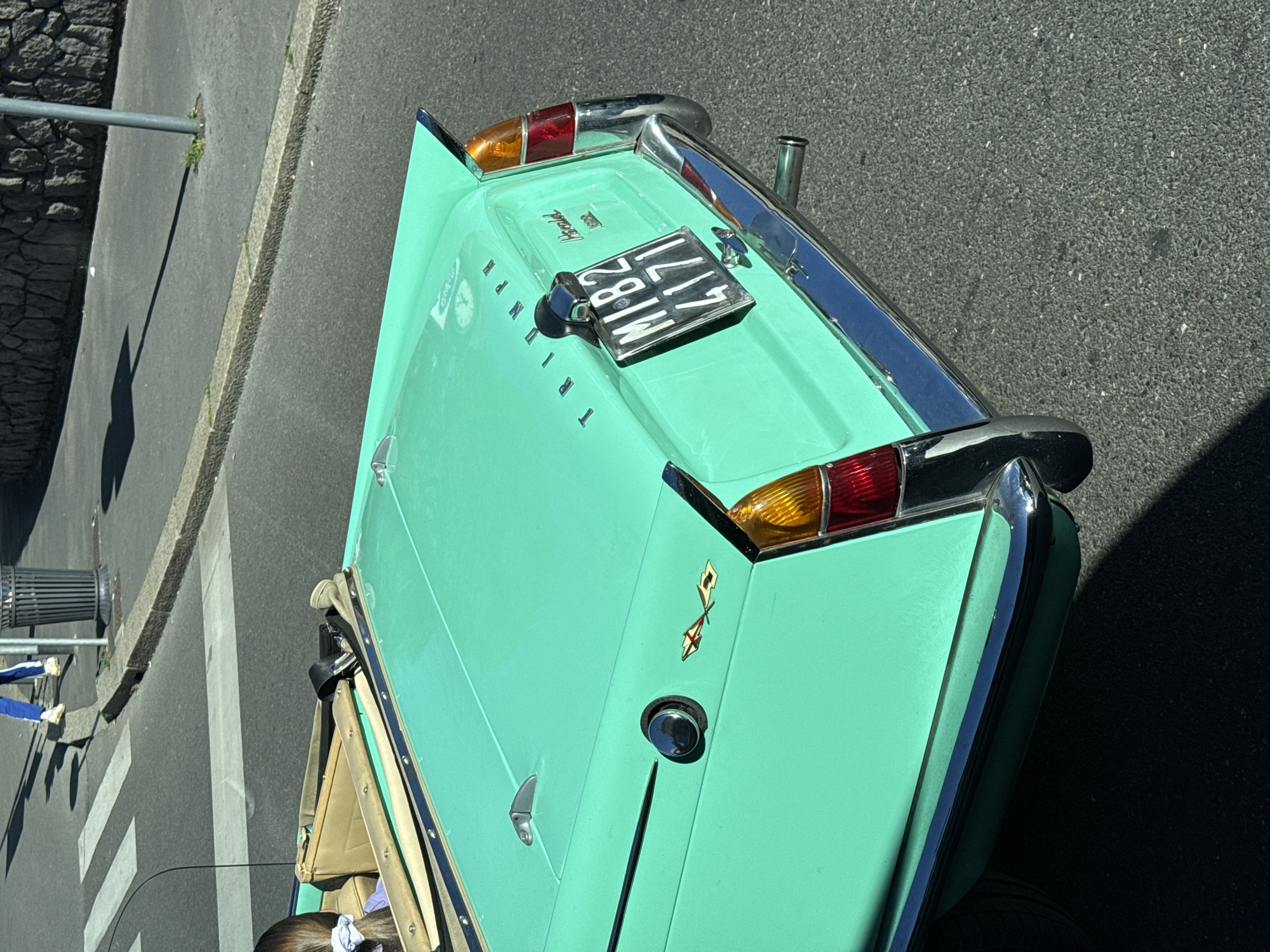 The image shows the rear of a light green Triumph Herald car parked on a street. The car has a chrome bumper and taillights. A black license plate reads "MI 82417" indicating it is a vehicle registered in Milan, Italy. The car is parked near a pedestrian crossing on a paved road. A curb and part of a stone wall are visible on the left side of the image. A person is visible inside the car in the lower left of the image. A small portion of a blue and white tracksuit is visible in the lower left as well as a trash receptacle.