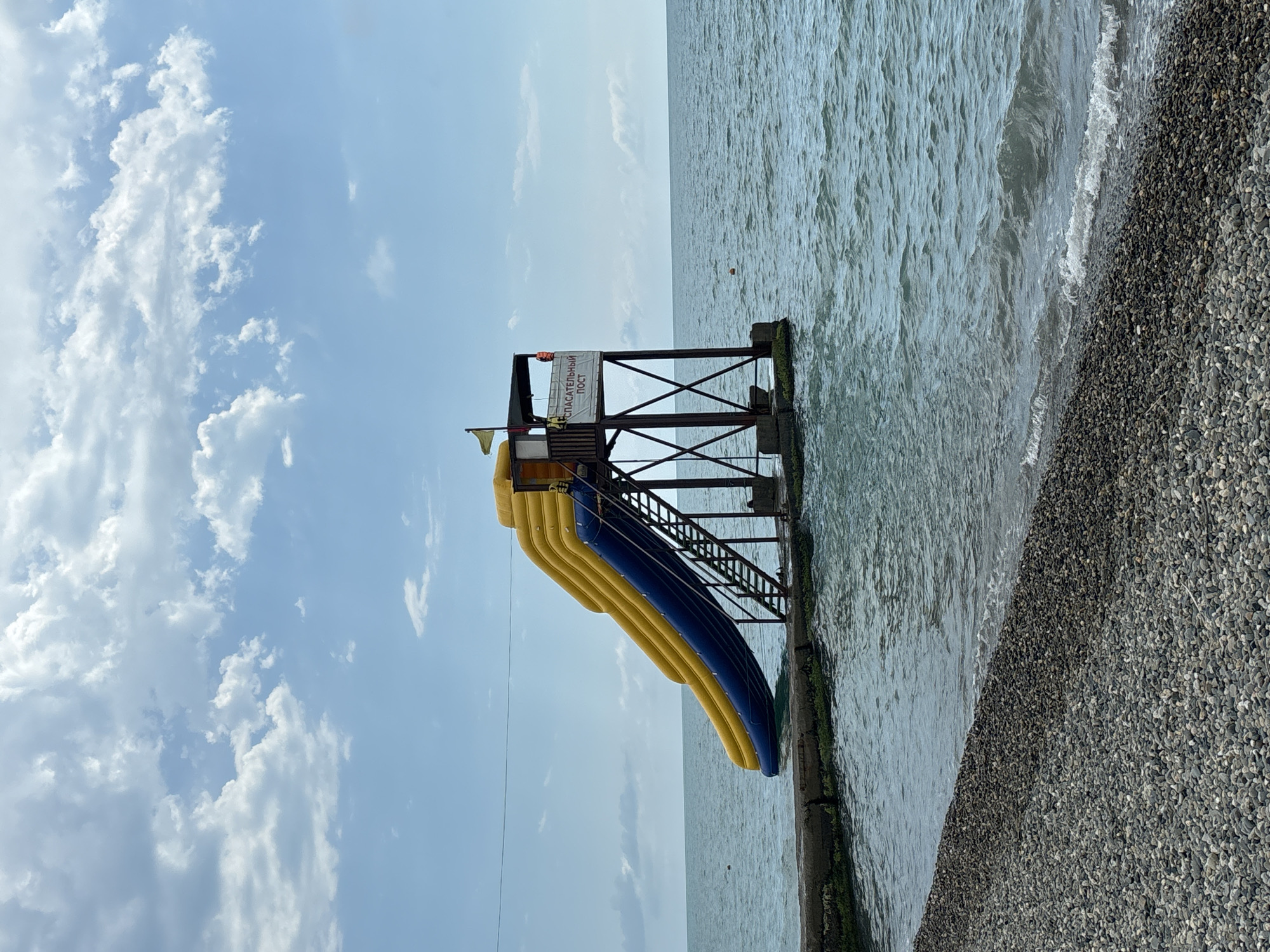 This media file depicts a view of the Black Sea coast in Sochi, Russia. The scene is dominated by a large, inflatable water slide, colored bright yellow and blue, mounted on a wooden structure extending over the water. The slide is positioned on a concrete embankment that runs along the shoreline, which is covered in dark pebbles. The sea is choppy with moderate waves breaking onto the shore, suggesting a breezy day.
Above the water slide, a lifeguard station is visible, marked with a white sign bearing text in Cyrillic which reads "СПАСАТЕЛЬНЫЙ ПОСТ" (SPASATEL'NYY POST), translating to "Lifeguard Post." A yellow flag is also flying from the station, commonly used to indicate safe swimming conditions.
The sky is a mix of light blue with scattered white cumulus clouds, indicating fair weather. The time of day appears to be afternoon, given the bright sunlight casting shadows. The overall atmosphere is one of a typical beach resort scene, likely during the summer season when such attractions are operational. There are no people visible in the frame, focusing the attention on the recreational infrastructure and the natural elements of the coast.