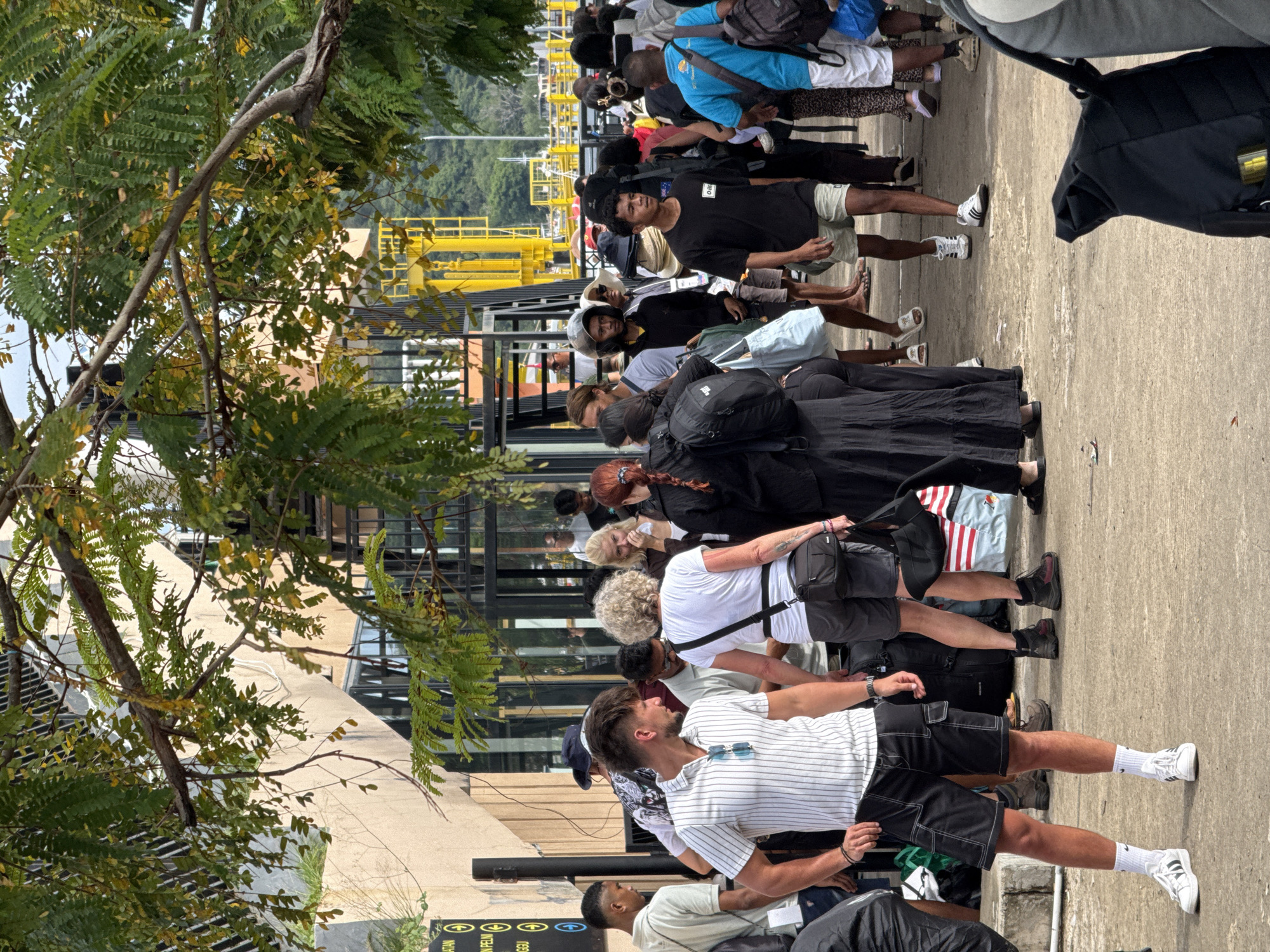 A medium-shot, medium-lit outdoor photograph taken at eye-level shows a line of people waiting to enter a building near the harbor in Labuan Bajo, Indonesia.
People of different ages and ethnicities are standing in a line with their backs facing the camera, looking in the same direction. Most of the people in line are wearing casual summer clothes and dark-colored backpacks. A few people are seen wearing masks.
The line of people is on a concrete surface, next to a building. There is a tree with green and yellow leaves on the left side of the frame. In the background, there is yellow harbor equipment with a body of water in the distance. There are more people in the distance to the right.