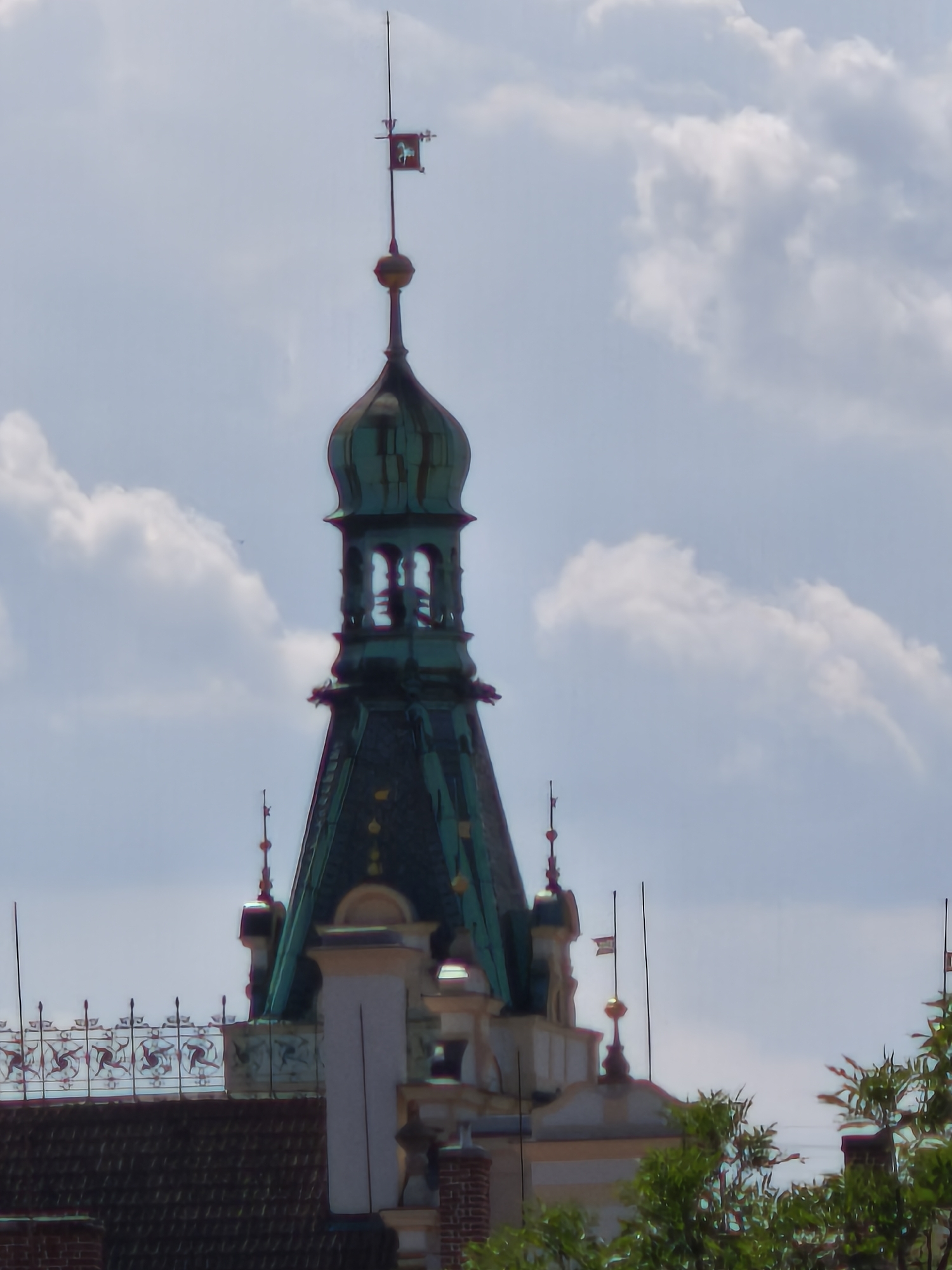 The image shows a detail of the tower of the Pardubice Castle in Pardubice, Czechia. The tower is topped with a weather vane in the shape of a horse. The sky is partly cloudy. The lower part of the image shows a portion of the castle's roof and surrounding buildings.