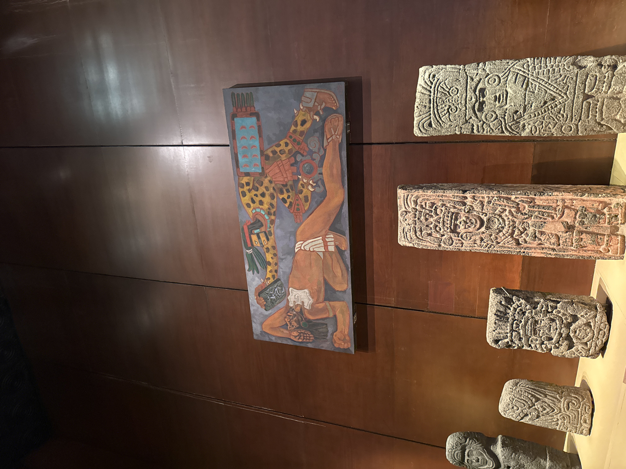 The image shows a portion of an exhibit in what appears to be a museum or cultural center, potentially located in Mexico City, Mexico, given the style of art. The background is a brown wall, likely made of wood panels. On the wall, there's a rectangular painting depicting a figure upside down, possibly related to Mesoamerican cosmology or mythology. The figure has markings reminiscent of a jaguar or similar feline, and is adorned with what might be ceremonial garb. To the right of the painting, there are several stone carvings or sculptures, rectangular in shape and stacked vertically. Each stone piece is adorned with intricate designs, possibly representing deities, symbols, or calendar systems from pre-Columbian cultures. The carvings are gray and have a weathered, aged appearance. The exhibit setting is professional, with the artifacts mounted securely on the wall.