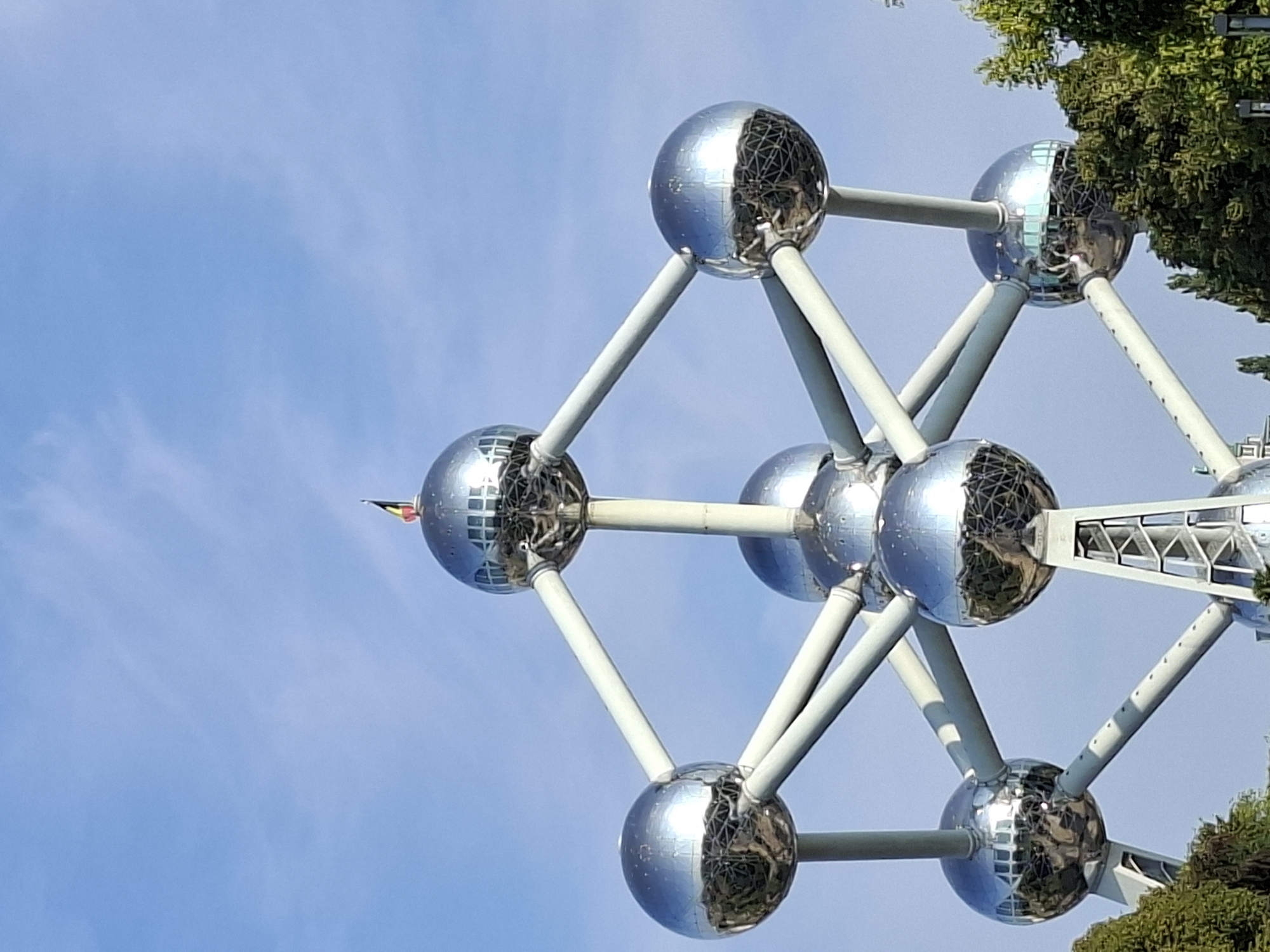 The image shows the Atomium, a landmark building in Brussels, Belgium, constructed for the 1958 Brussels World's Fair. The structure consists of nine spheres, each representing an atom, connected by tubes. In the foreground, the iconic Atomium structure is prominently displayed against a clear blue sky. A small Belgian flag is visible atop the uppermost sphere. The reflective surfaces of the spheres capture the surrounding environment, including trees and sky. The lower portion of the image shows the tops of lush green trees, indicating an outdoor setting. The Atomium is illuminated by daylight, suggesting it is daytime.