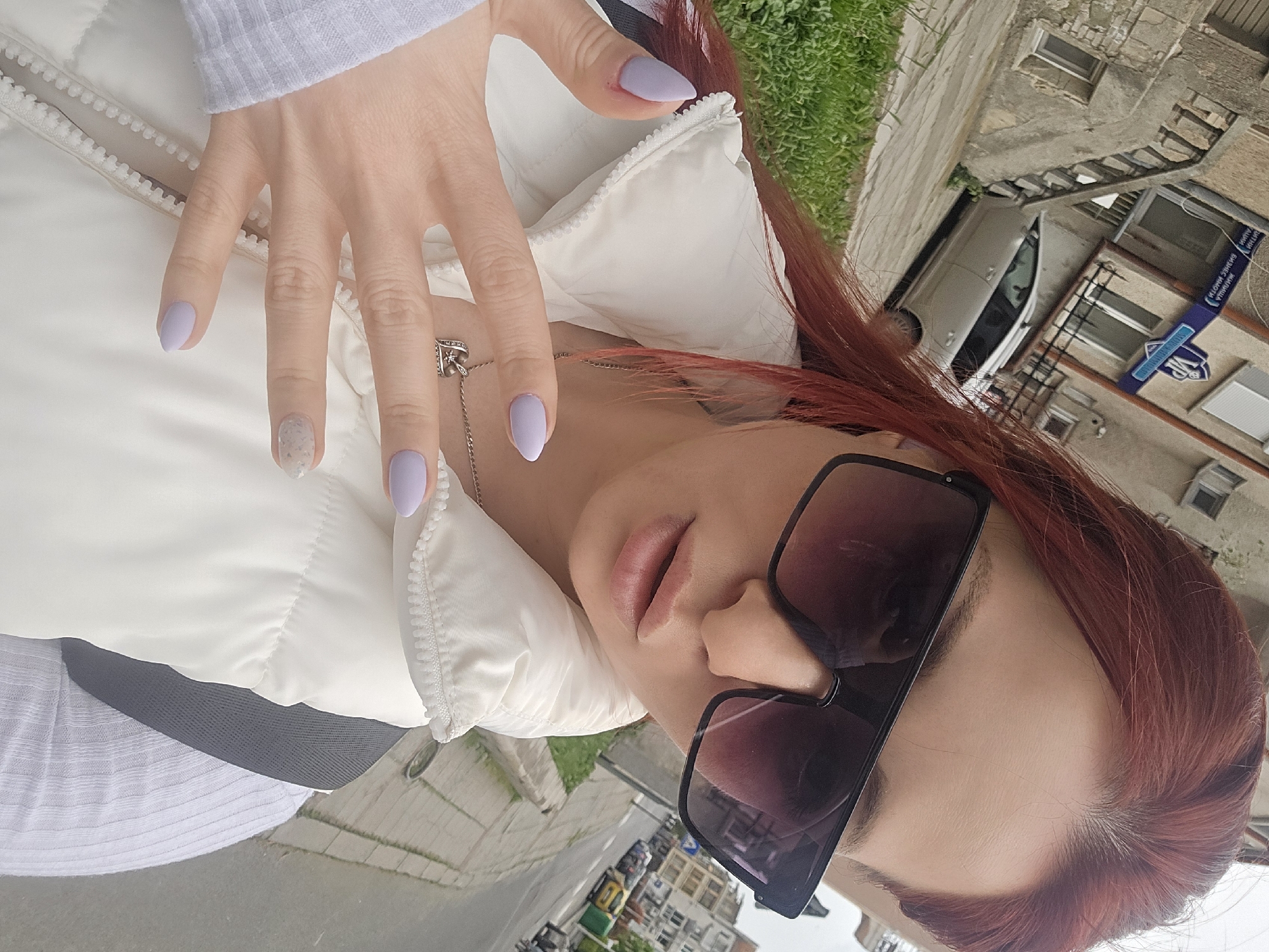 A woman with reddish-brown hair and sunglasses is wearing a white vest. She has light purple nail polish, one with a glitter effect. She's wearing a silver necklace with a heart-shaped pendant. The background shows a street scene. A silver car is parked near a building with a blue and white sign indicating "VIP". Based on the Cyrillic text on the sign and the general architectural style, the location is likely in Burgas, Bulgaria. The overall scene appears to be an outdoor shot taken during daylight.