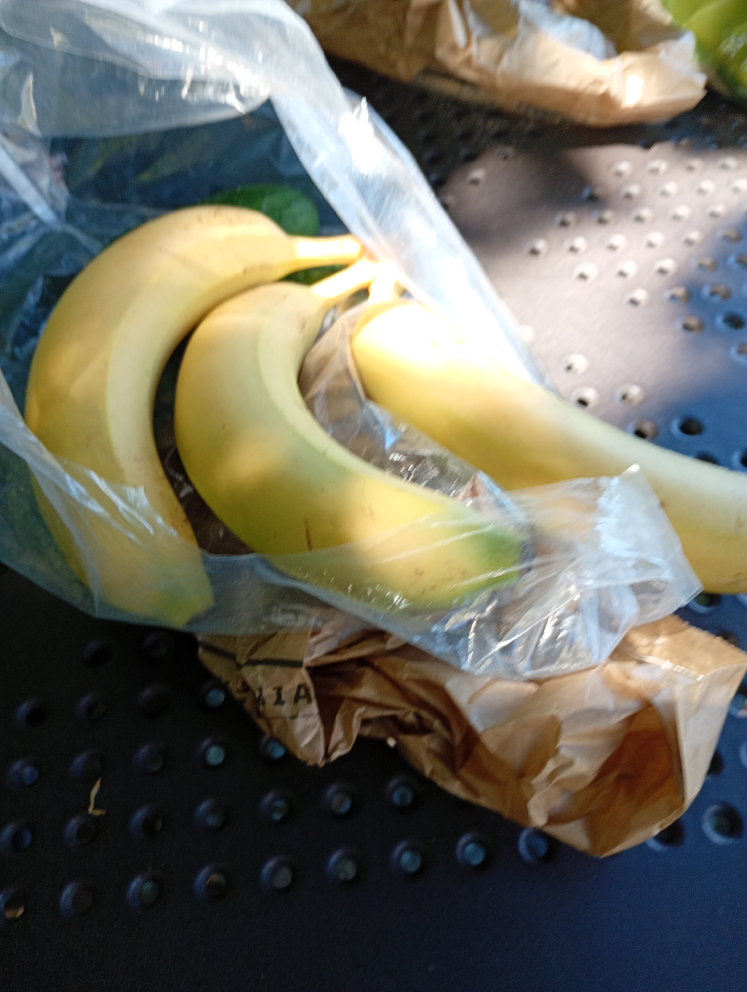 A close-up shot shows three bananas inside a clear plastic bag. The bananas are yellow with some green at the tips, indicating they are not fully ripe. The plastic bag is slightly crinkled, and some of the bananas are nestled in a brown paper bag beneath the plastic. The background is a dark, textured surface, possibly a table, with a pattern of raised circular dots. The lighting suggests an outdoor setting, possibly in the city of Międzyrzecz, Poland, given the provided context. No people are visible in the image.