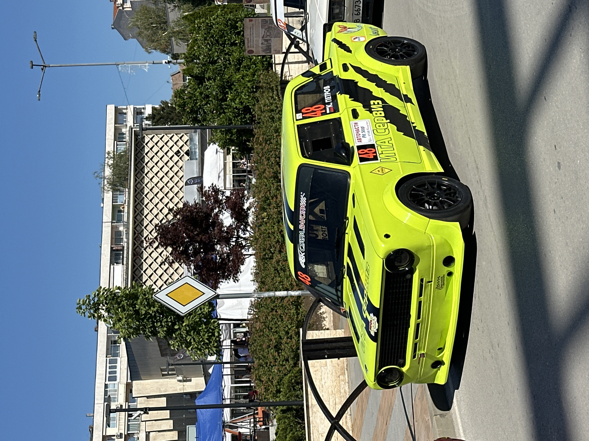 An eye-level, medium shot captures a brightly colored, neon yellow race car parked on a paved street in Gorna Oryahovitsa, Bulgaria. The car, adorned with black racing stripes and decals, dominates the foreground. Visible text on the car includes "48", "Авточасти Pit Stop", and "ИТА Сервиз". The car's front bumper is modified with additional lights. Behind the car, a low, decorative metal fence lines the sidewalk. Beyond the fence, a well-maintained green hedge separates the sidewalk from a paved area where several white tents and blue canopies are set up. People are visible milling around behind the hedge, under the tents. In the background, a multi-story building with a distinctive geometric facade stands tall. A diamond-shaped road sign with a yellow center and white border is mounted on a pole near the hedge. The sky above is a clear, bright blue, and sunlight casts shadows on the street. To the right of the yellow race car, a portion of another white car with a visible number "48" and text "Б. ПЕТРОВ" is also visible.