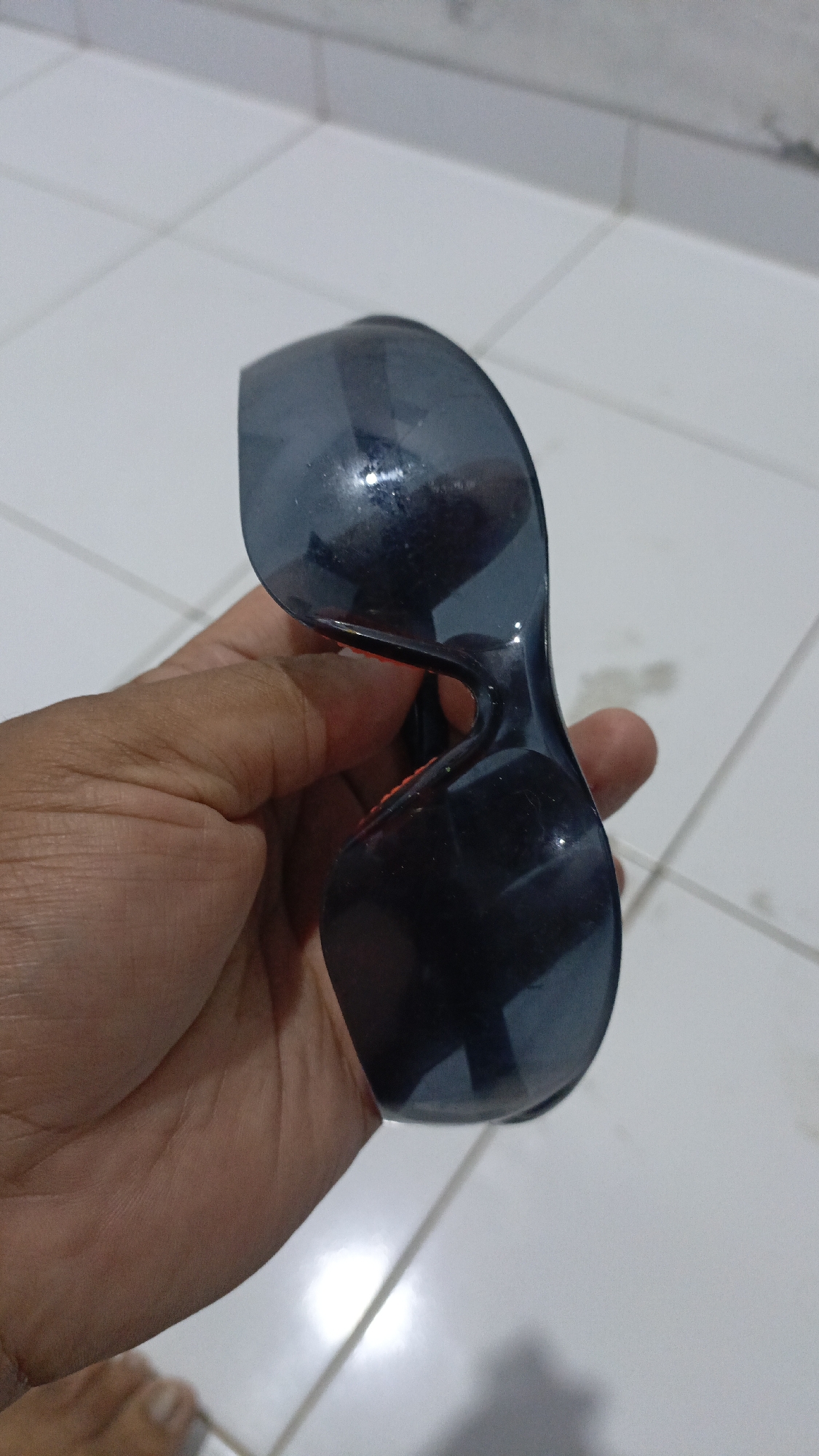 A hand holds a pair of dark, wraparound sunglasses against a tiled background. The sunglasses have a thick, black frame and dark lenses, reflecting the surroundings. The tiled surface appears to be a floor, with visible grout lines and a slightly worn appearance. The lighting in the scene is bright, creating some glare on the sunglasses. The background is a light-colored tiled floor, likely in Trihanggo, Indonesia.