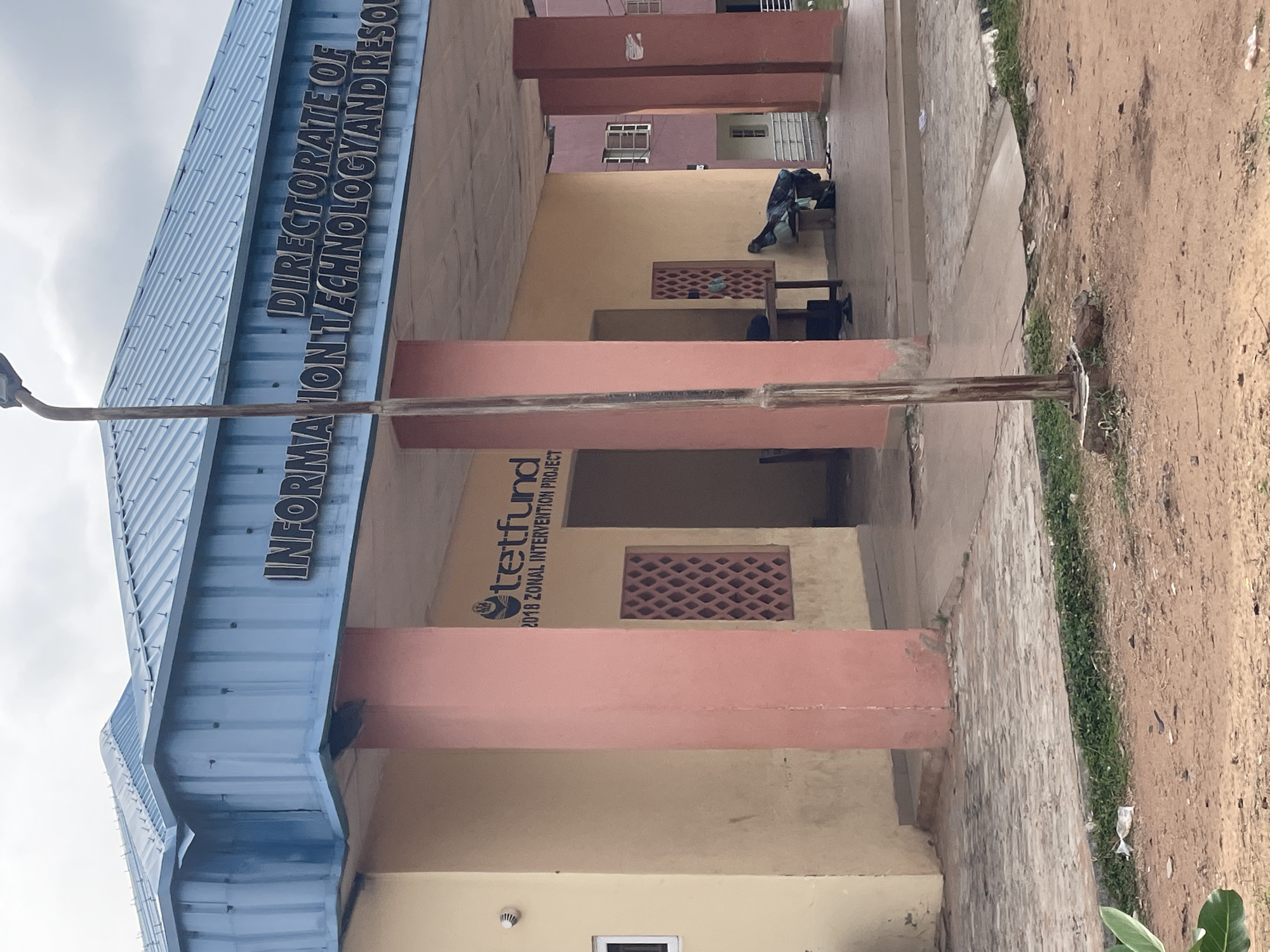 The image shows the "DIRECTORATE OF INFORMATION TECHNOLOGY AND RESOURCE CENTER" building in Lokoja, Nigeria. The building has a blue corrugated metal roof and light yellow and pink painted walls. Large pink pillars support the roof over an open-air walkway. The walkway has a brick-paved floor with some debris scattered on it. A sign on the wall reads "tetfund 2018 ZONAL INTERVENTION PROJECT." Inside the walkway, there are two tables and some bags of trash. The background shows a multi-story pink building with windows. A lamppost stands in front of the building. The ground in front of the walkway is dirt with some sparse green vegetation.