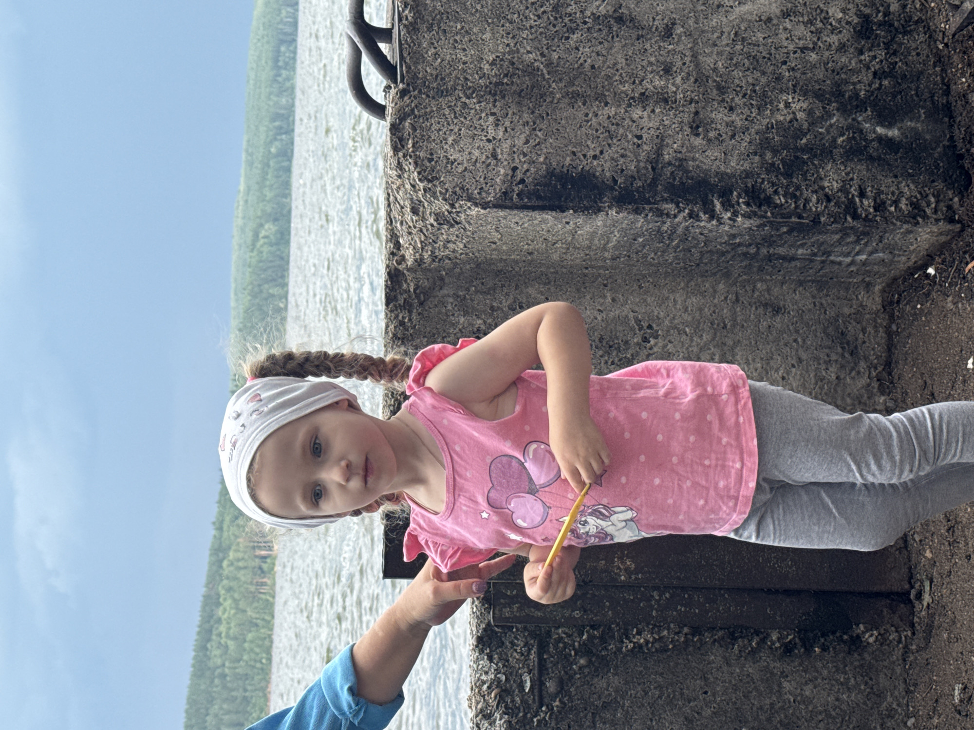 A young girl with blonde braided hair, wearing a white headscarf, a pink t-shirt with a unicorn and heart design, and grey leggings stands on a concrete embankment near a body of water. An adult's arm, wearing a blue shirt, is visible on the left side of the frame, with their hand gently resting on the girl's shoulder. The girl holds a yellow object, possibly a crayon or a thin stick, in her hands. The background features a wide, rippling body of water, with a densely forested shoreline and a cloudy sky above. The scene appears to be located along the Kama River embankment in Chaikovsky, Russia, known for its picturesque views and recreational areas.