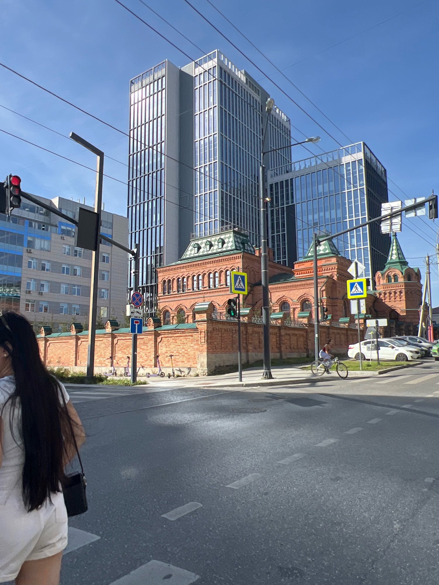 In Yekaterinburg, Russia, on a sunny day, the scene captures a street view with modern architecture and historical elements. A tall, glass-fronted building stands prominently, reflecting the blue sky and surrounding structures. Adjacent to it is a lower, historic building constructed of red brick with distinctive green accents on its roofline and towers. A red brick wall with a decorative top encloses the historic building.
In the foreground, a person with long, dark hair, wearing a white top and white shorts, is walking away from the camera, carrying a black crossbody bag. The street in front of them has white crosswalk markings. To the right, a cyclist is riding a bicycle across the street. Several parked scooters are visible behind the red brick wall.
Traffic lights and street signs are present, indicating an intersection. Cars are parked on the right side of the road, and another vehicle is driving. The sky is clear and blue. Overhead, power lines and streetlights are visible. The overall environment suggests an urban setting with a mix of old and new urban development.