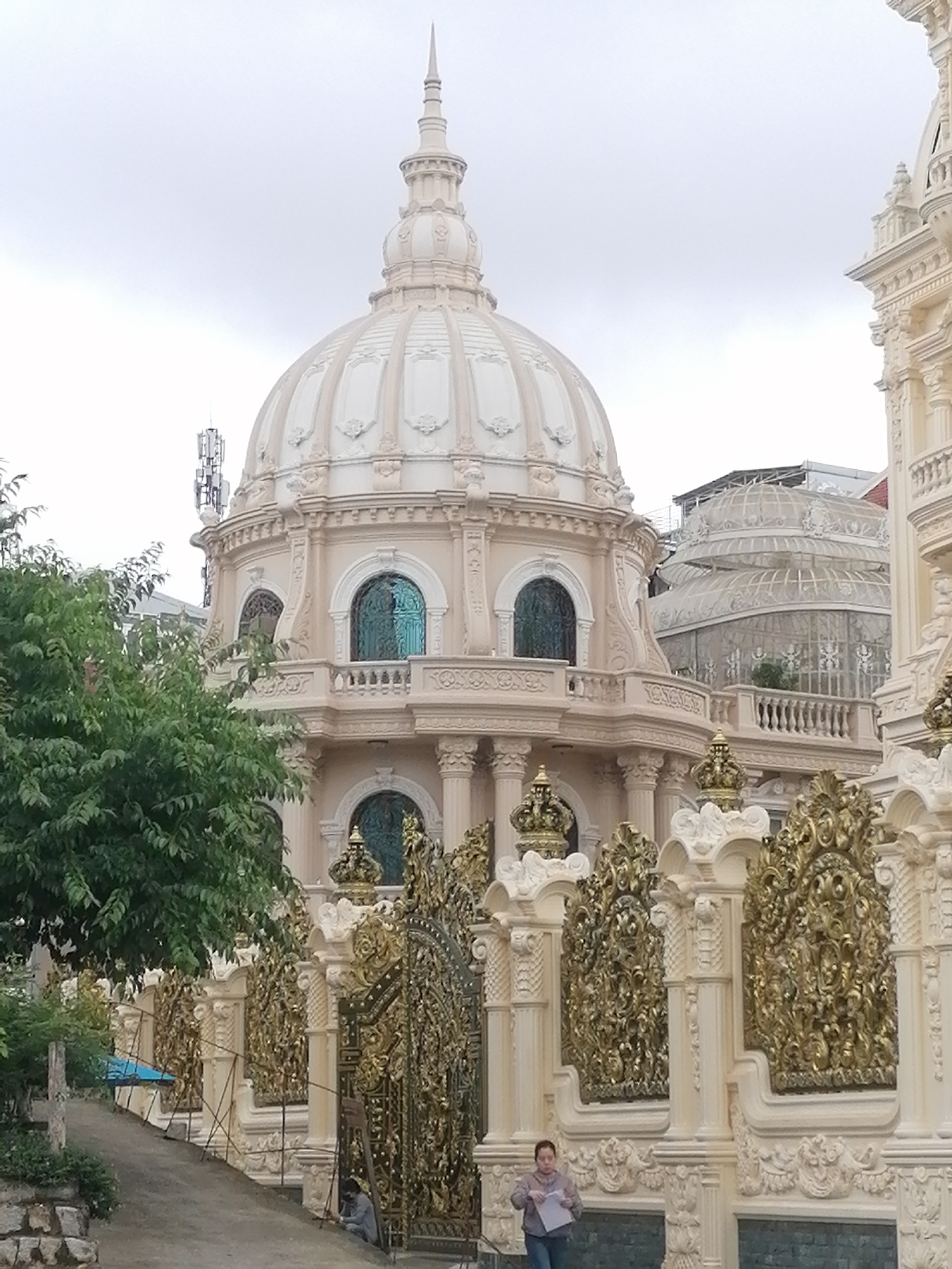 This media file captures a grand, ornate building under an overcast sky. The central feature is a large, light-colored (beige or cream) structure topped with a prominent dome and a spired finial. The dome is segmented and adorned with intricate decorative moldings. Below it, the building features several levels with arched windows, some of which appear to have distinct turquoise-colored glass or grilles. Balustrades, columns, and elaborate architectural details are visible throughout the facade. A cellular antenna tower is situated behind the dome on the left.
To the right, another section of an equally elaborate building is partly visible, also featuring white pillars and ornate carvings. In the background, behind the main domed structure, a large glass greenhouse-like building with a decorative roof is discernible.
Separating the buildings from a paved pathway in the foreground is an elaborate fence or gate system. This fence comprises numerous white pillars topped with crown-like elements and intricate golden, foliage-like panels.
On the paved pathway in the lower-right, a woman wearing a light-colored top and jeans stands, looking down at papers she holds. Further along the pathway, near one of the golden gates, another person is seen crouched, possibly engaged in work. Lush green trees and foliage frame the left side of the scene. The architecture suggests a luxurious and highly decorative style, possibly in Da Lat, Vietnam, given the context. No visible text is identifiable.