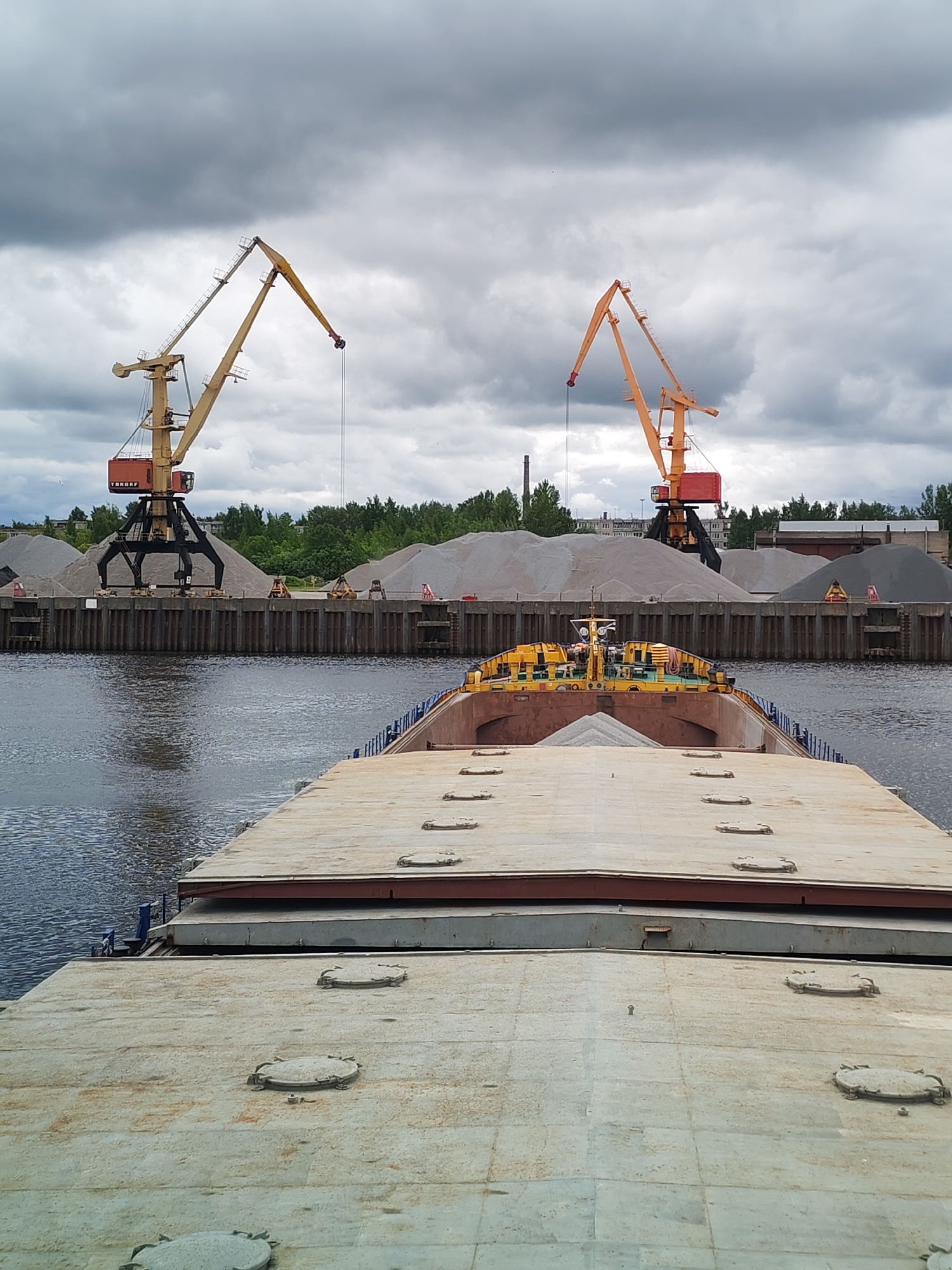 The image depicts a barge loaded with gravel in what appears to be a port or industrial area in Rybinsk, Russia. In the background are two large cranes, each with a different color scheme (one yellow and black, the other orange and black), situated on a quay with stockpiles of gravel. The foreground is dominated by the barge's deck, showing various circular hatches and a section containing a load of gravel. The river fills the midground, reflecting the overcast sky above.