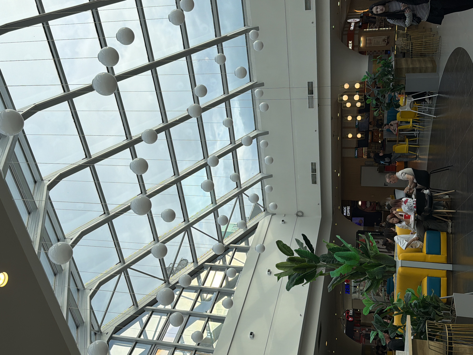 The image depicts the interior of a modern shopping mall, likely a food court area, with a prominent glass ceiling that allows natural light to filter in. A large number of spherical white lights are suspended from thin wires, creating a decorative ceiling feature. Below the glass ceiling, a large indoor plant with broad green leaves adds a touch of nature to the space.
In the background, there are various commercial establishments, including a sign that reads "MAGIC COFFEE". Several tables and chairs are arranged in the seating area, with some people seated and appearing to be eating or conversing. One table has yellow chairs and a yellow booth with blue accents. A woman with long dark hair is seated at one of the tables, wearing a white outfit and a shirt with a character print. Another person, possibly a man, is seated at the same table, wearing a white shirt. Further back, another seating area is visible with more people.
The overall atmosphere seems casual and relaxed, suggesting a typical day in a public space. The time of day is not explicitly indicated, but the natural light suggests it is daytime. The weather appears to be overcast, as seen through the glass ceiling. The presence of multiple food vendors and seating areas clearly identifies this as a food court or a similar dining space within a mall. The overall aesthetic is modern and bright, with a mix of geometric structures and organic elements from the plant.