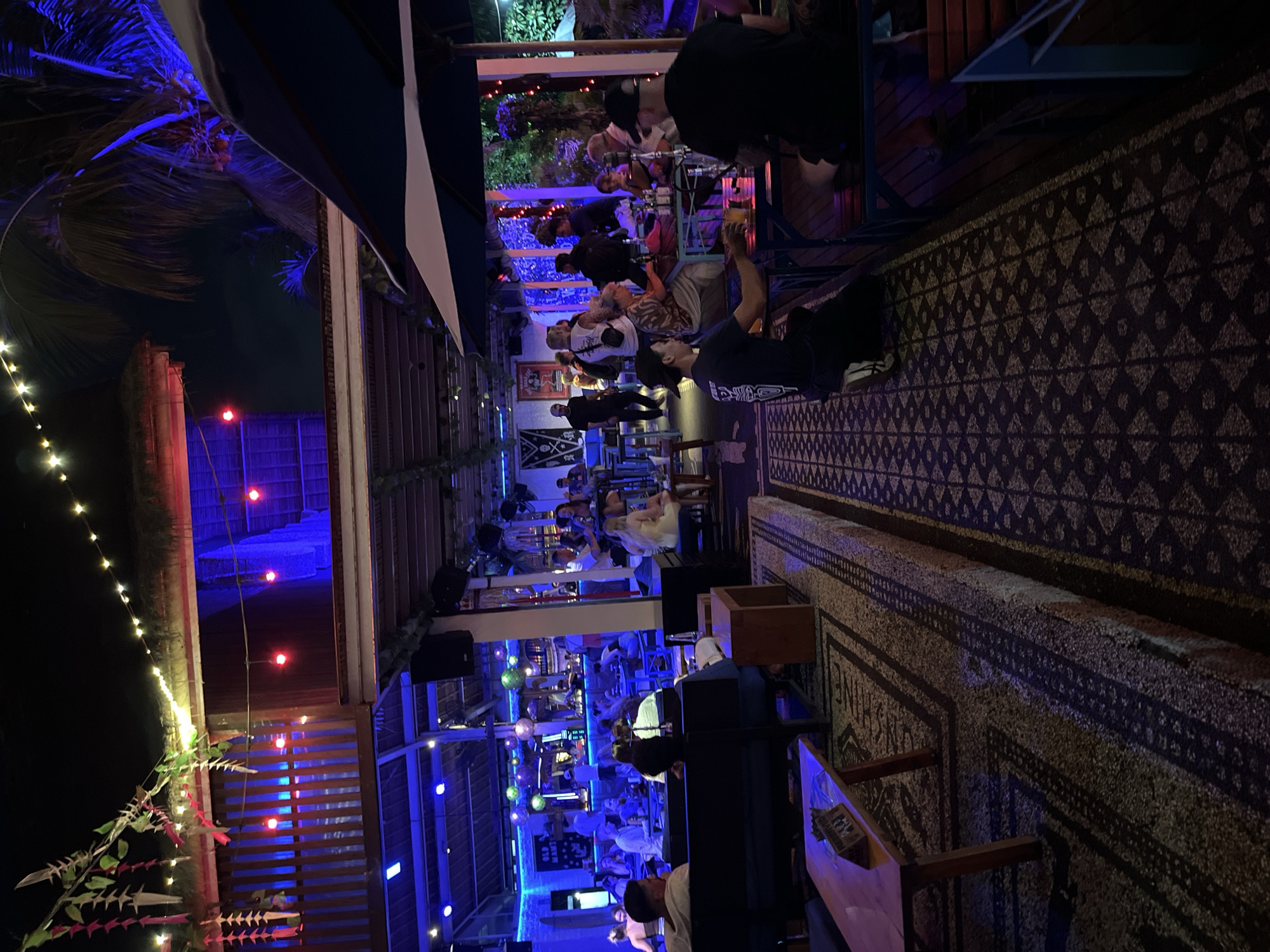 A night scene at a crowded open-air establishment in North Kuta, Indonesia. The establishment appears to be a bar or restaurant, with a lively atmosphere. The lighting is predominantly blue and red, creating a vibrant, party-like ambiance. Several people are seated at tables, some with drinks, and others are standing or mingling. The architecture suggests a tropical setting, with open-air sections and thatched roofs visible. String lights are draped across the ceiling, adding to the festive mood. The floor is covered with patterned rugs, and the furniture consists of dark wooden tables and seating. The overall impression is that of a popular nightlife venue.