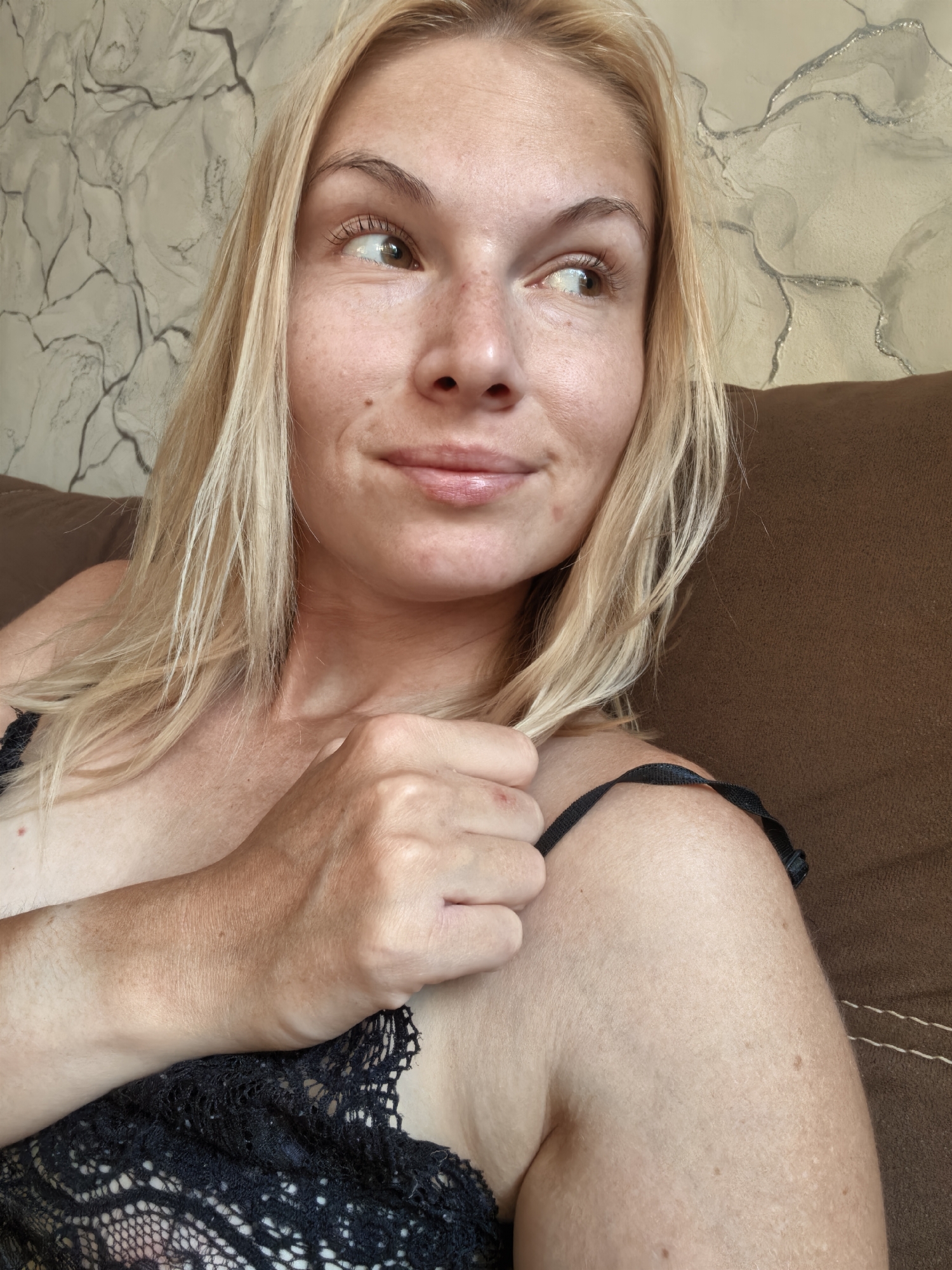 A blonde woman with fair skin and freckles is shown in a close-up portrait. She is wearing a black lace top and is looking to her right. Her left hand is resting on her shoulder, and her hair is partially obscuring her face. The background appears to be a textured wall with a brown couch. The image is undated and its location is unknown, with no indications of Sterlitamak, Russia.