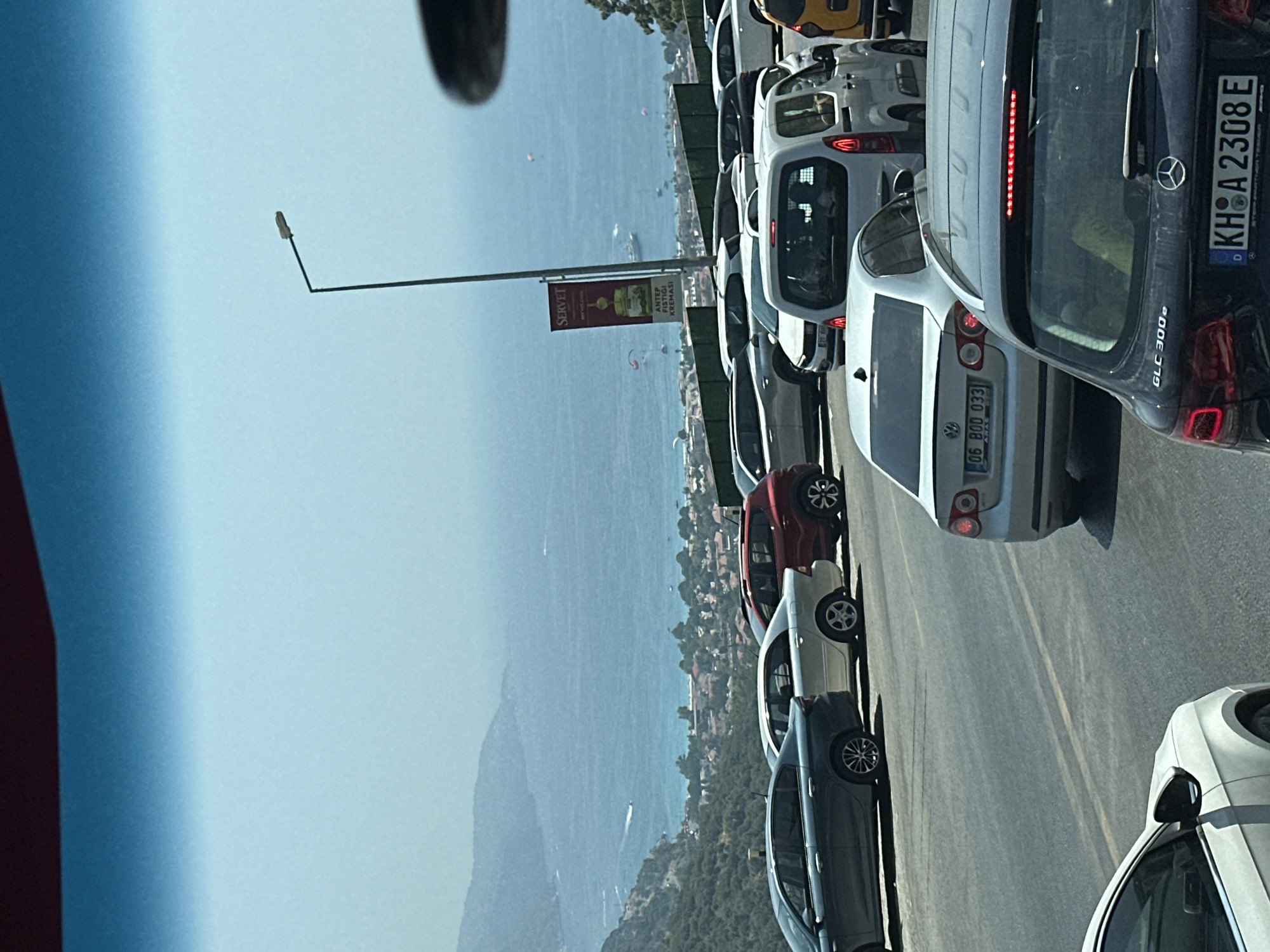 The image shows a traffic jam on a road overlooking the sea and the city of Fethiye, Turkey. The road is on a hillside, with cars lined up as far as the eye can see. In the background, the blue sea stretches out to the horizon, dotted with boats and possibly paragliders. The city of Fethiye is visible on the coast, with its characteristic red-tiled roofs. The sky is hazy and blue, suggesting a warm, sunny day. The cars are of various makes and models, and the license plates are visible on some of them. The scene suggests a popular tourist destination, likely experiencing peak season traffic.
