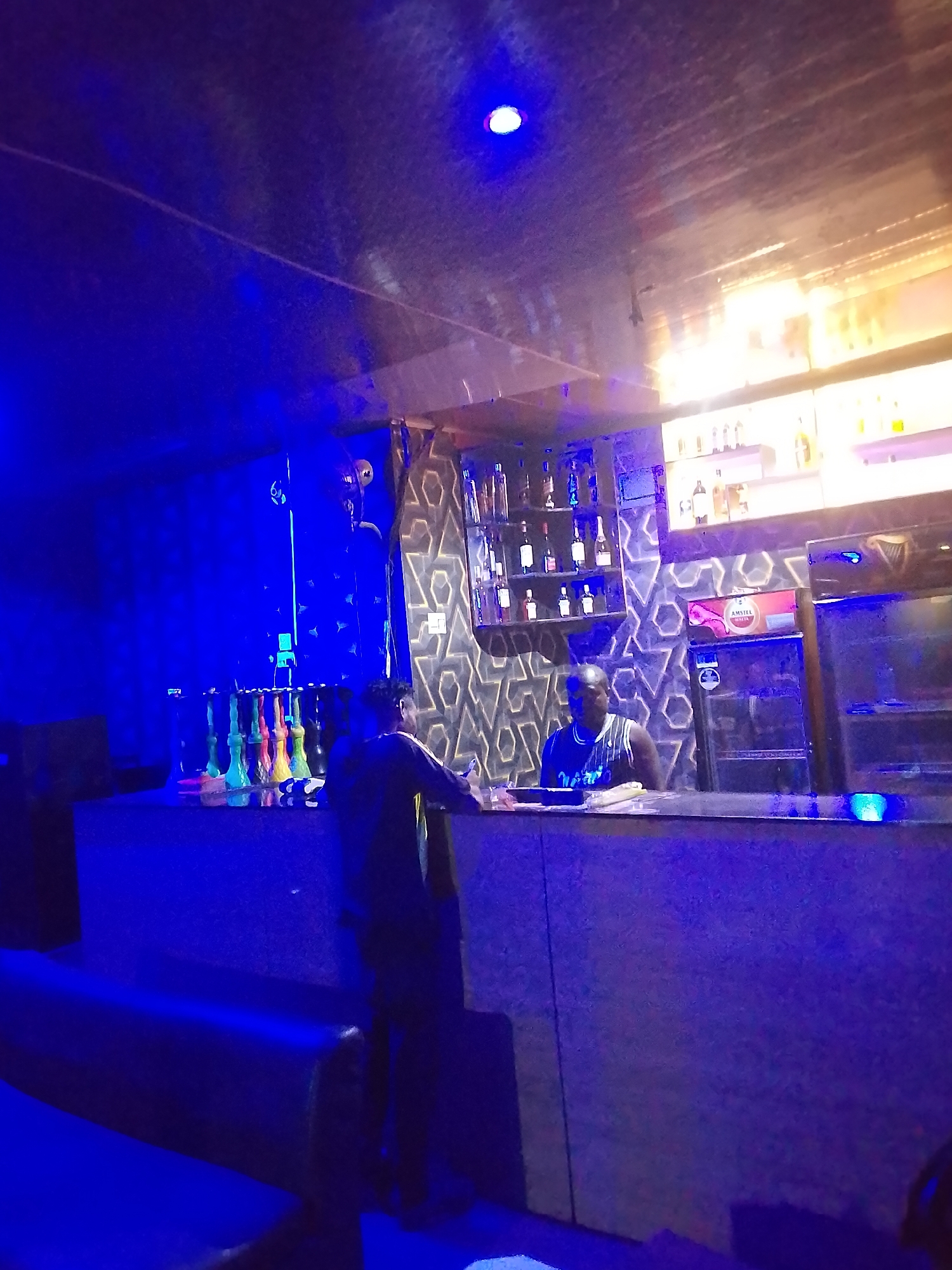 This image depicts an indoor bar or lounge at night, characterized by its prominent blue artificial lighting. Two men are present: a bartender, wearing a dark sleeveless top, stands behind a dark counter, and a customer, dressed in a dark long-sleeved shirt, stands in front of the counter with his back mostly to the viewer. They appear to be engaged in an interaction at the bar.
Behind the bartender, the wall is adorned with a decorative geometric pattern. Liquor bottles are displayed on shelves above the counter, and two refrigerated units are visible to the right, one of which has a red box with the text "AMSTEL MALTA" clearly visible. On the left side of the counter, a collection of colorful shisha or hookah pipes is arranged. A bright blue light fixture is visible on the dark ceiling. The foreground shows hints of dark seating or benches for patrons. The overall setting suggests a casual, modern social venue.