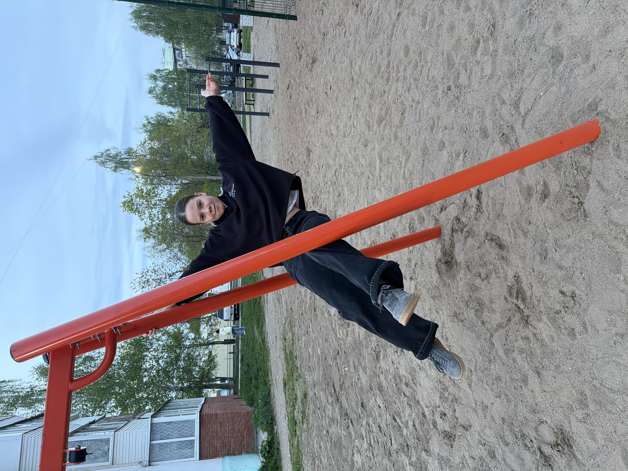 A person, presumably a young adult, is hanging on a red-painted metal climbing structure in what appears to be a playground or recreational area. The person is wearing a black hoodie and dark-colored jeans, with gray sneakers. They are facing the camera, with one arm raised. The ground beneath the structure is covered in sand. In the background, there are trees, a building (possibly an apartment complex), and other playground equipment. The overall scene suggests an urban environment. The location is the city of Chaikovsky, Russia.