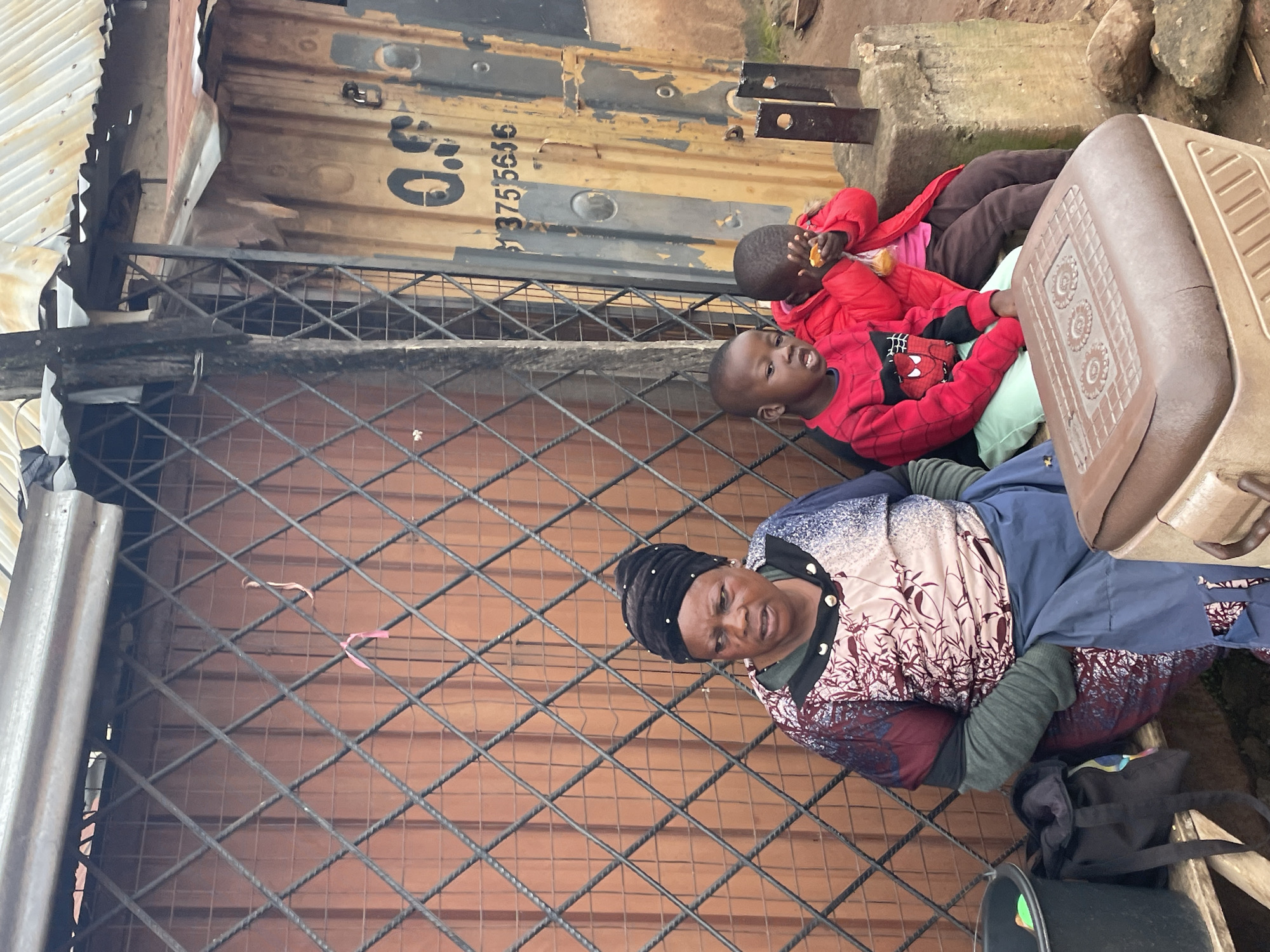 An outdoor scene in Jos, Nigeria, shows an adult woman and two young children sitting on a bench in front of a metal gate. The woman wears a black headscarf and a patterned dress. The children are dressed in bright red jackets. The child on the right is eating. Behind them is a corrugated metal structure with yellow paint and some black lettering. A cooler sits in the foreground. The ground is dirt and small stones are visible.