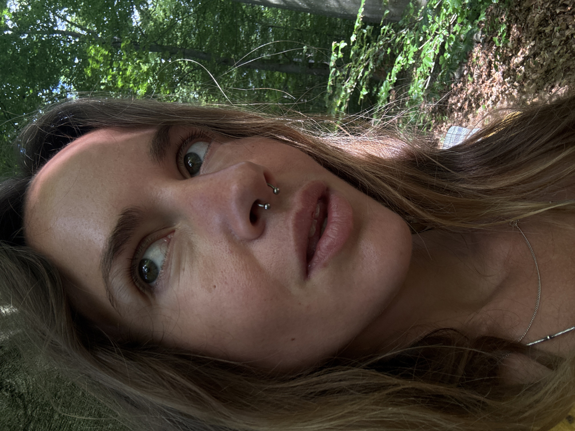 A close-up, outdoor, chest-up shot focuses on a young adult female's face, turned to the right. Her eyes are looking upward and to the right. She has light brown hair, parted on the left, with some strands falling across her face. She has a septum piercing with a silver horseshoe-shaped ring. Her lips are slightly parted, revealing some teeth. She is wearing a thin silver chain necklace. The background is a forest with green foliage and dappled sunlight on the ground. The scene is outdoors in an unspecified natural environment.