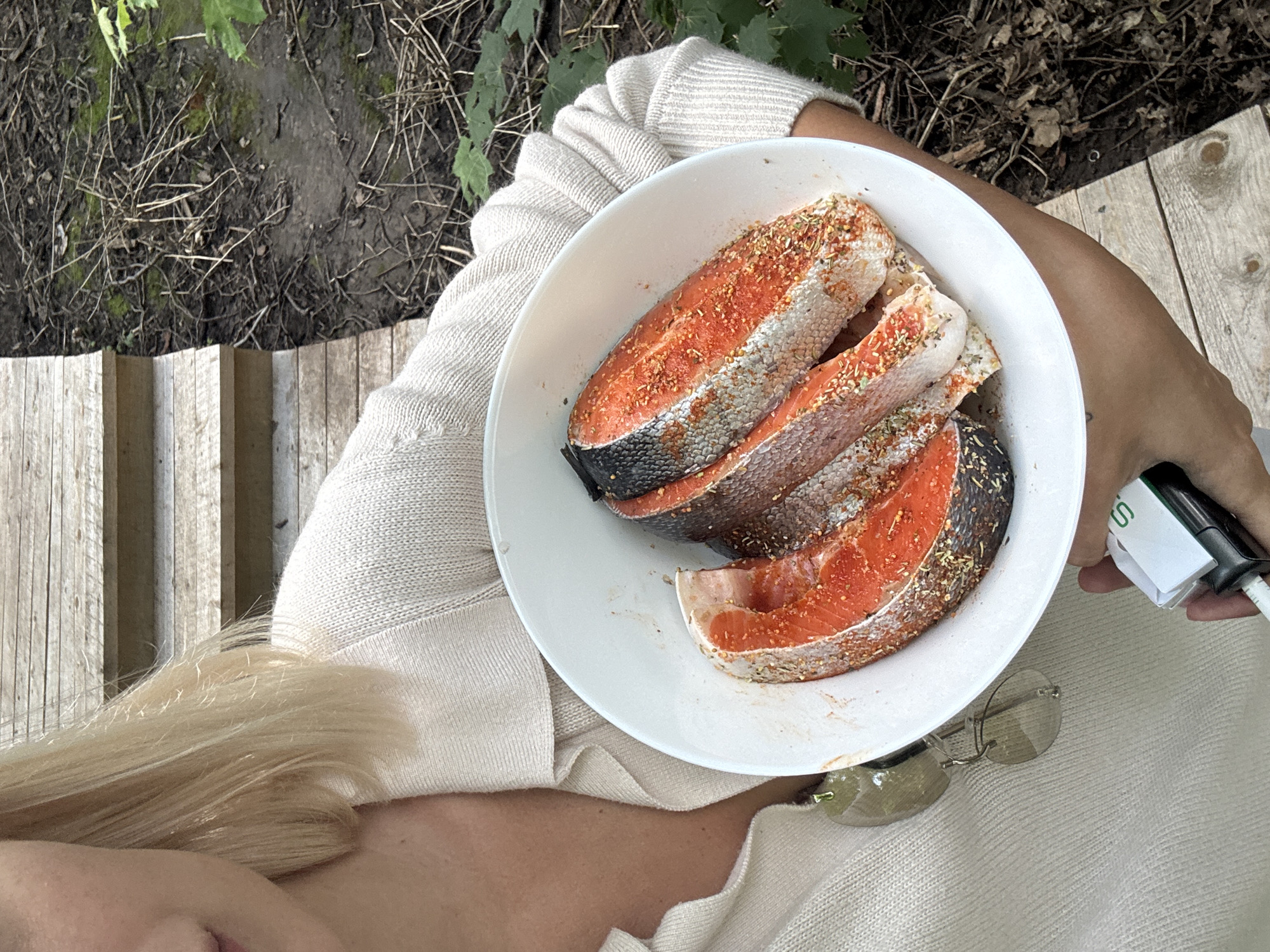 A blonde woman, partially visible from the chest up, holds a white bowl containing three seasoned salmon steaks. The woman wears a light-colored knitted cardigan. She stands outdoors on a wooden walkway, with a natural, somewhat overgrown area to her right. The ground beside the walkway is covered in dirt, dried leaves, and twigs. In her right hand, she holds a small white object with a green "S" logo, possibly a vape or a similar device. Her left arm is not visible. The scene appears to be in a natural setting outside of a city.