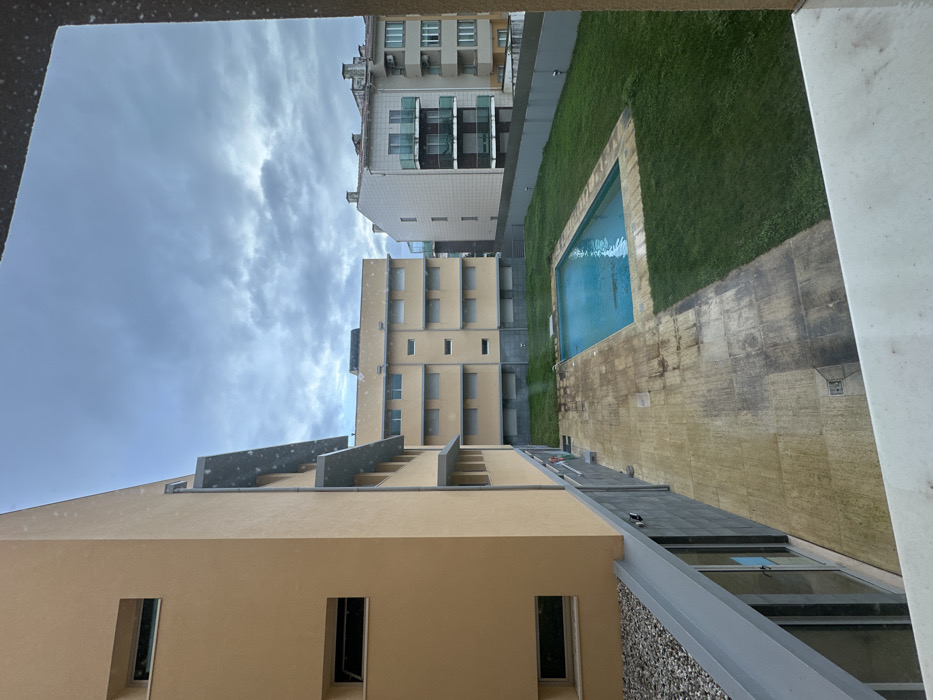 The image displays an urban courtyard in Gafanha da Nazaré, Portugal, viewed from an elevated vantage point under an overcast sky. To the left, a multi-story building with a light beige facade features rectangular windows, some open, and gray architectural overhangs. Its lower section has a dark gray tiled base with ground-level glass doors and an adjacent patch of gray gravel. The courtyard's center contains a rectangular swimming pool with clear blue water, bordered by light-colored, discolored pavers exhibiting stains. Green grass surrounds the pool and paved area. In the background, directly opposite the pool, a second multi-story building with a light beige facade and numerous shuttered windows is visible. To its right, a white-tiled residential building featuring multiple dark-framed balconies with glass railings is partially visible, with another building further in the distance. A low, dark gray wall defines the courtyard's right boundary. The scene is devoid of people or active interactions.