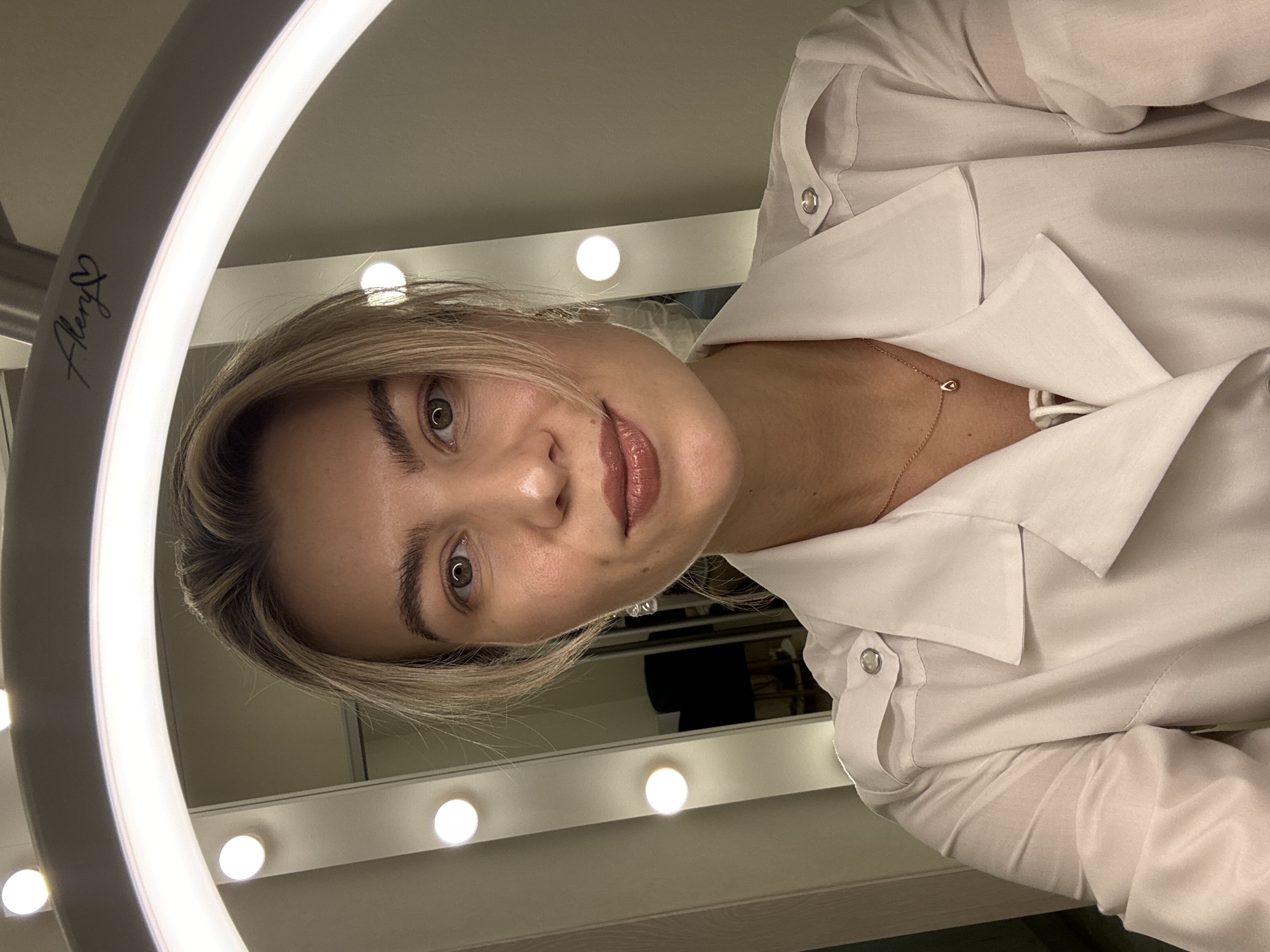 A young woman with blonde hair and light skin is looking at the camera. She is wearing a white collared shirt and a gold necklace with a small pendant. Her expression is neutral. The woman is in front of a ring light, with a mirror and vanity lights visible behind her. The location appears to be indoors, likely a dressing room or a similar space. The specific city is unknown.
