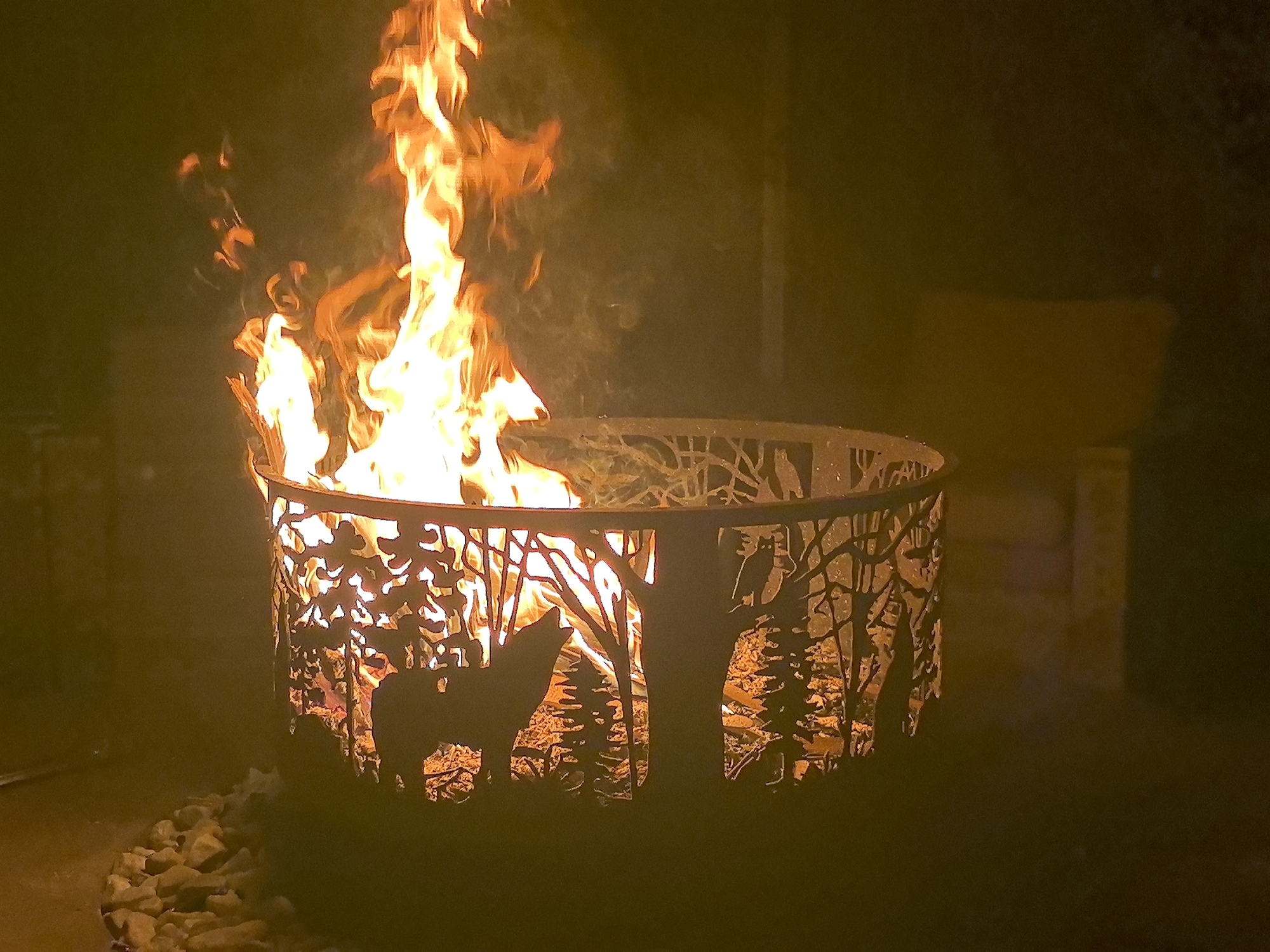 An outdoor fire pit at night, illuminated by a bright, upward-reaching flame. The fire pit is cylindrical and made of metal, with intricate cutouts depicting a forest scene with a wolf howling at the moon and an owl perched on a branch. The fire burns within the pit, casting an orange glow through the cutouts and illuminating the surrounding area with warm light. In the foreground, a scattering of light-colored stones surrounds the base of the fire pit. The background is dark and indistinct, suggesting a nighttime setting. The location is specified as the city of Taldykorgan, Kazakhstan.