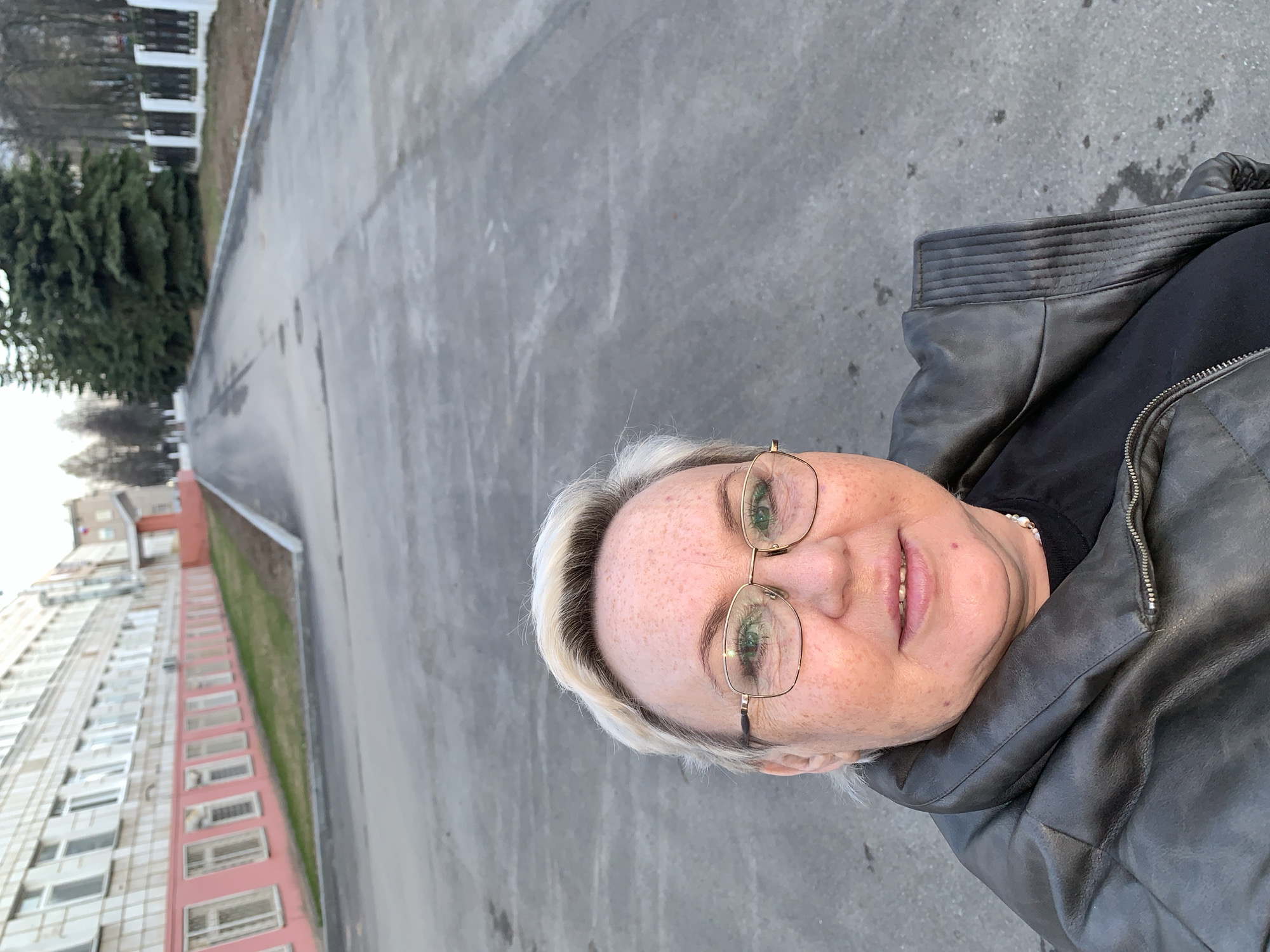 A woman with light blonde hair and glasses takes a close-up selfie outdoors in Chaikovsky, Russia. She is smiling gently at the camera, revealing freckles on her face and green eyes behind gold-rimmed spectacles. She wears a dark grey or black leather jacket with a ribbed collar and a visible zipper over a black shirt, with a string of small pearls visible around her neck.
The setting appears to be an urban or suburban environment. Behind her, a wide, grey asphalt or concrete surface stretches into the background, possibly a road or a spacious path. To the left, a long, multi-story building with numerous windows and a distinctive pink or red lower section extends along the side of the path. Further in the background, green coniferous trees and white fences or railings are visible under a bright, overcast sky. The overall lighting suggests daytime, possibly late afternoon or morning, with soft, diffused light and no harsh shadows. There is no visible text in the image.