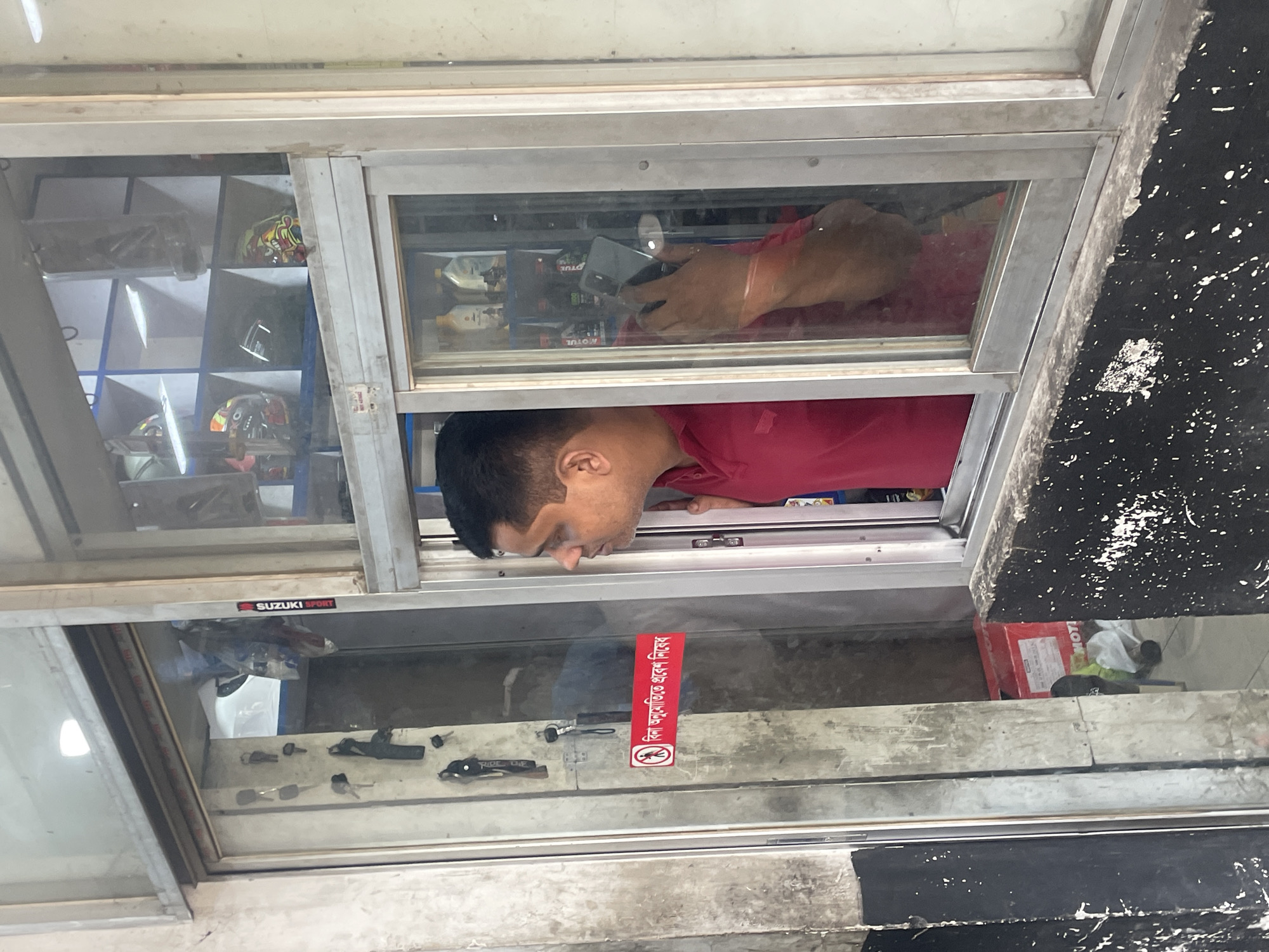 A man wearing a red shirt is seen from the chest up behind a glass window in a shop in Dhaka, Bangladesh. He is looking down and to the left. In the window above him, an individual is using a smartphone. On the shelves behind the glass are motorcycle parts and accessories, and a sticker on the window reads "No helmet, no service" in Bengali. The shop displays a "SUZUKI SPORT" logo. A red box labeled "MOTUL" is visible in the lower right corner of the frame. The outside of the building appears to be weathered and dirty.