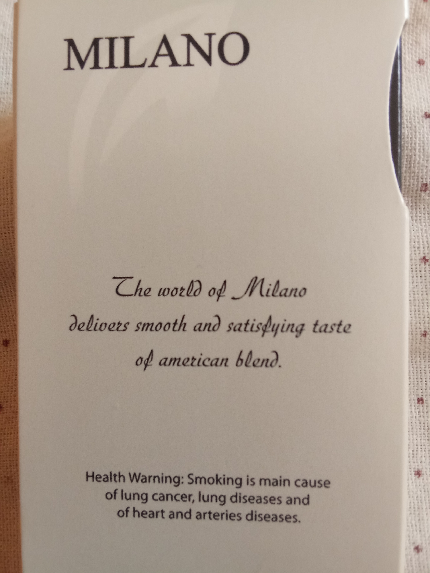 The image shows the front of a cigarette pack. The brand name "MILANO" is printed in large, dark, sans-serif font at the top. Below this, in a smaller, cursive font, are the words "The world of Milano delivers smooth and satisfying taste of american blend." At the bottom of the pack, in a standard font, is a health warning: "Health Warning: Smoking is main cause of lung cancer, lung diseases and of heart and arteries diseases." The background of the cigarette pack is white, and the pack itself is shown against a textured, light-colored fabric with small red circular patterns. There are no people or any indication of a specific location in Fergana City, Uzbekistan.