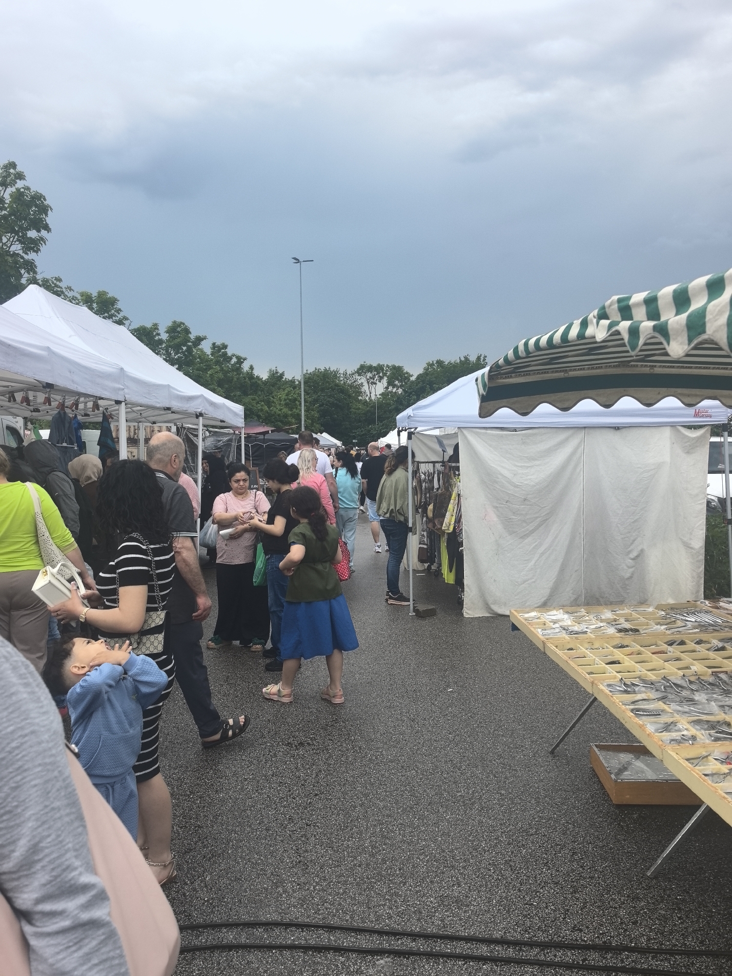 The image depicts an outdoor market in Wallau, Germany. Several white tents, some with striped canopies, are set up on what appears to be a paved surface, likely a parking lot. People of various ages and ethnicities are browsing the vendors' offerings. One table displays small items in divided sections. The sky is overcast, suggesting recent or impending rain. There is a light post in the background. Electrical cables are visible on the ground in the foreground.