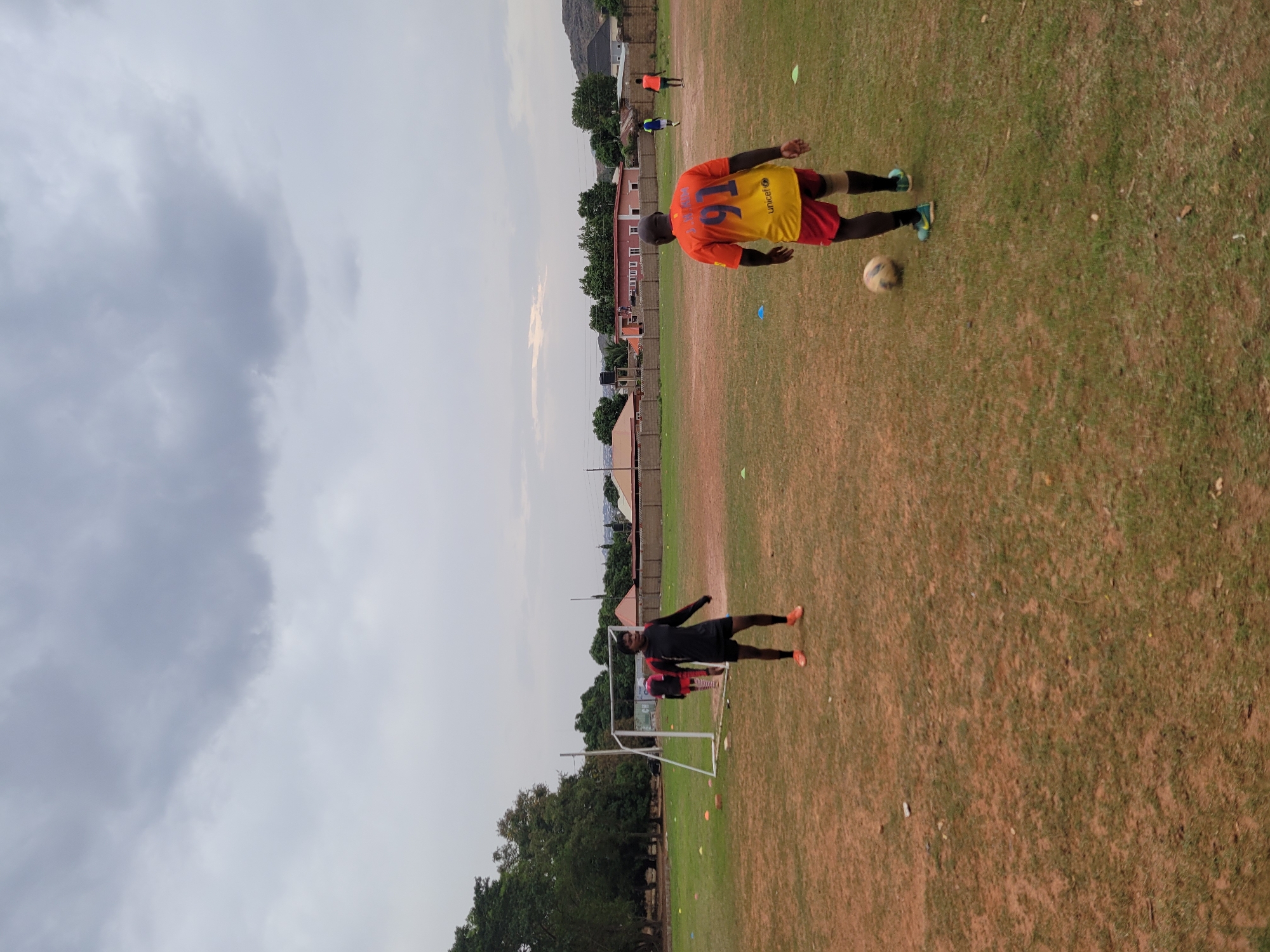 On an outdoor field in Jos, Nigeria, under a cloudy sky, two individuals are engaged in a soccer-related activity. One person, wearing an orange and yellow jersey with the number 16, is kicking a soccer ball. Another individual, dressed in black with red accents, is positioned near a soccer goal. Cones are scattered on the field, and buildings and trees are visible in the background.