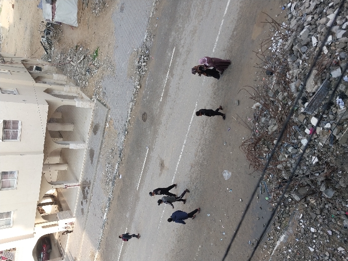 An elevated photograph captures a street scene in Khan Yunis, Palestinian Territory. The immediate foreground is dominated by a substantial pile of concrete rubble, twisted rebar, and assorted debris, partially obscured by several dark utility cables crossing diagonally. A grey asphalt road occupies the midground, featuring two faded white longitudinal markings and scattered small debris. Seven individuals are walking on the road: a group of four, predominantly male and dressed in dark clothing (one wearing a blue jacket), moves from left to right; separately, a male in dark clothing walks from left to right, passing two females in long garments and head coverings (one black, one light purple) who are walking from right to left. In the background, a multi-story, light-colored building with an arched, open ground-floor arcade stands alongside the road. To the building's left, a red car is visible under a covered area. To the building's right, an unpaved area of dirt and rubble extends to a makeshift white tent structure and further scattered debris.