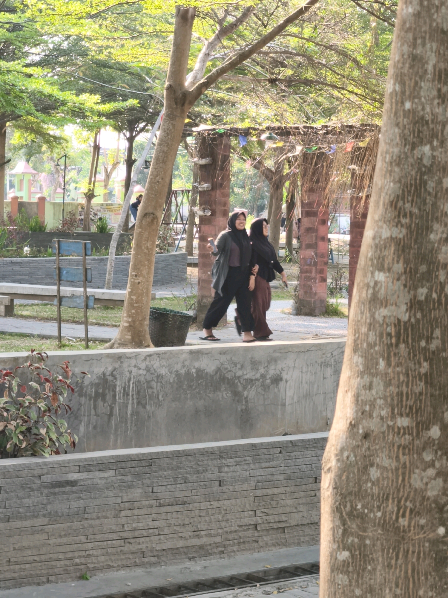 Two young women, both wearing hijabs, are walking side-by-side on a raised concrete path in a park setting. The woman on the left is wearing a dark grey cardigan over a dark purple top and black pants, with her left arm extended as if gesturing. The woman on the right is wearing a black hijab and a dark brown long-sleeved top with brown pants. They are walking towards the right side of the frame. The path they are on overlooks a lower level area with stone wall construction. In the background, there are trees with lush green foliage, some brick structures that appear to be part of an open-air pavilion decorated with colorful flags, and a tall tree trunk in the foreground on the right. The scene suggests a sunny day in a park located in Desa Dibal, Indonesia, as indicated by background signage that is partially visible and reads "KOTA PERTIWI."
