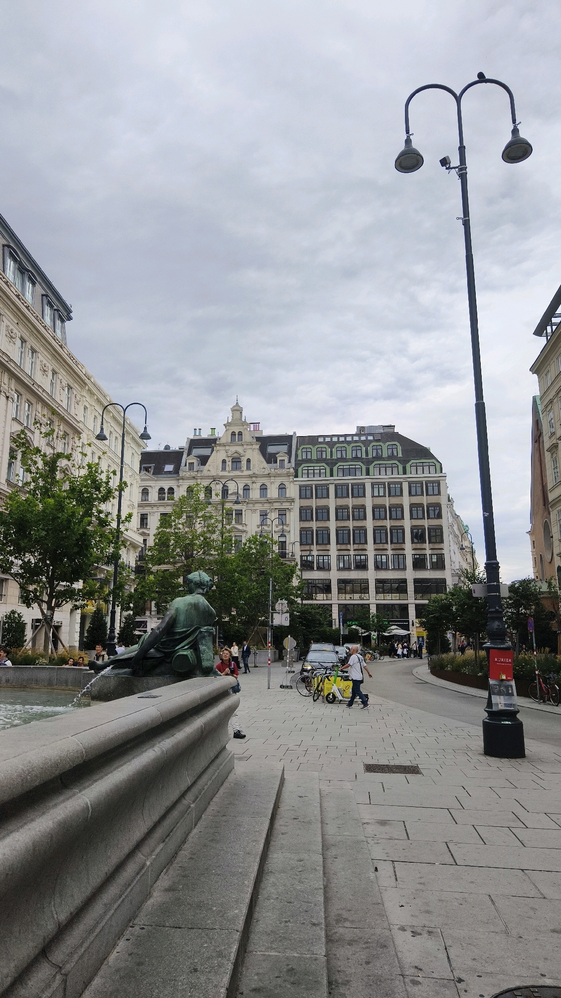 The image captures a street scene in Vienna, Austria, with a focus on the architecture and public space. In the foreground, a stone fountain structure is visible, featuring a bronze statue of a reclining figure. Beyond the fountain, a wide, paved street lined with trees leads toward several multi-story buildings. The buildings exhibit characteristic Viennese architecture with ornate facades and mansard roofs, one prominently featuring a green roof. A double-headed lamppost stands tall on the right side of the street, adorned with a sign reading "KURIER." Several people are present, some walking and others gathered, indicating a public square or thoroughfare. Yellow public bicycles are parked on the right side of the street, and a car is stopped in traffic. The sky is overcast with clouds. The overall impression is a typical urban setting in Vienna, showcasing its blend of historical architecture and modern city life.