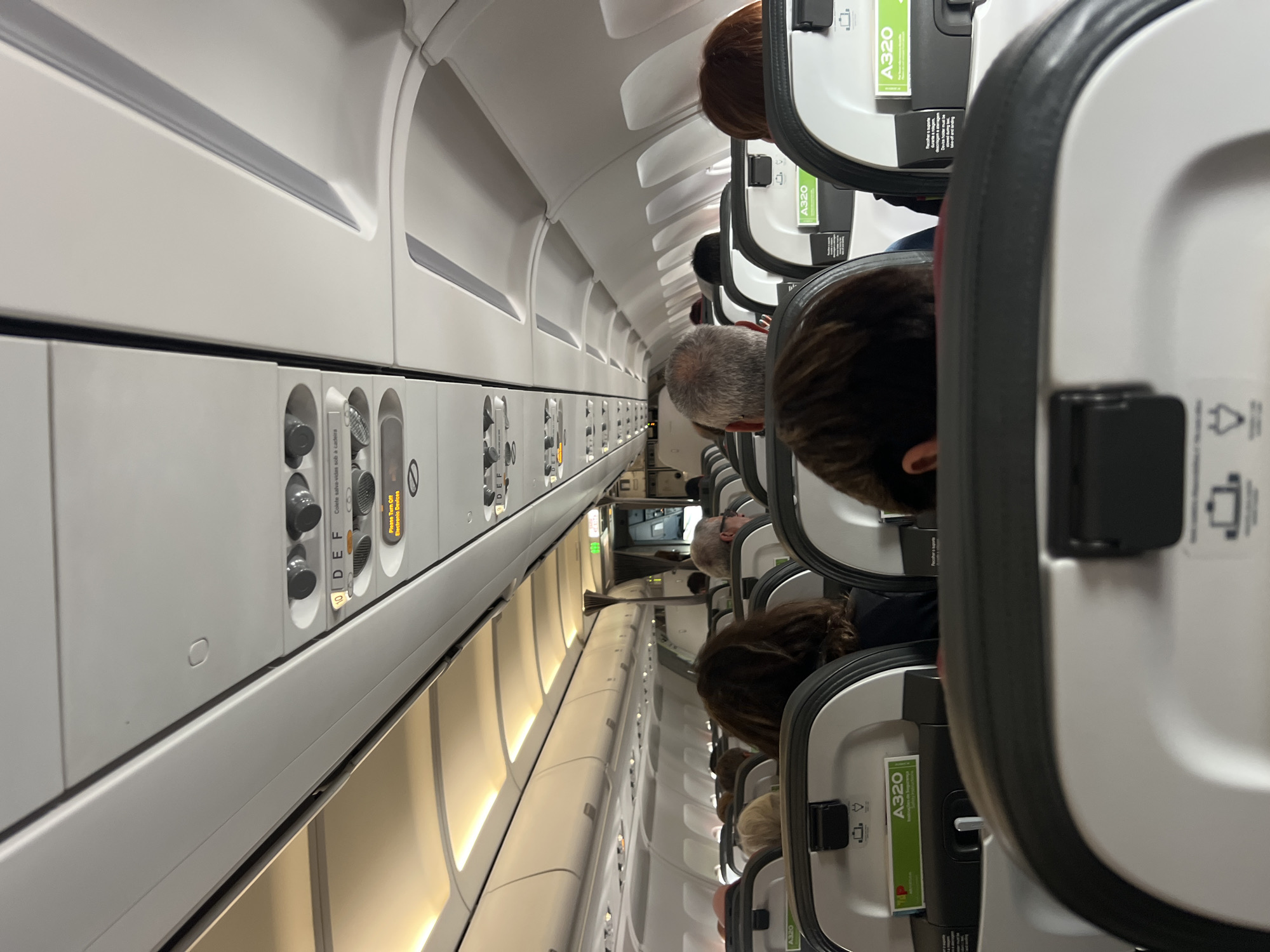 The image shows the interior of an airplane cabin, likely at Paris-Orly Airport in Paray-Vieille-Poste, France, given the context of an aircraft. The perspective is from a passenger's seat, looking forward down the aisle. Multiple rows of seats are visible, occupied by passengers, their heads and backs of their heads are the primary subjects. The overhead compartments and lighting fixtures are prominent in the upper portion of the frame, indicating a typical commercial airliner interior. Safety information cards are visible in seat pockets. The overall environment suggests a pre-flight or mid-flight scene within an airplane.