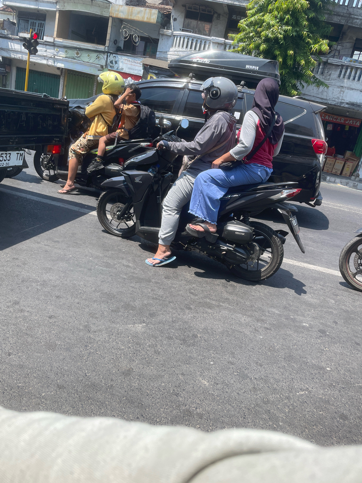In a street scene in Jogosari, Indonesia, two people are riding on a dark-colored scooter. The person in front is wearing a grey long-sleeved shirt, grey pants, and a grey helmet. The person behind is wearing a pink shirt, a dark hijab, and blue pants. Both are wearing flip-flops. The scooter is in motion, indicated by the blur of the wheels. Another scooter is partially visible to the right, also in motion. In the background, a black SUV with a roof carrier is stopped in traffic. To the left, the back of a dark pickup truck is visible, with a license plate reading "533 TM". A traffic light with a red signal is mounted on a pole to the far left. Buildings line the street, with some trees and awnings visible. The road surface is asphalt.