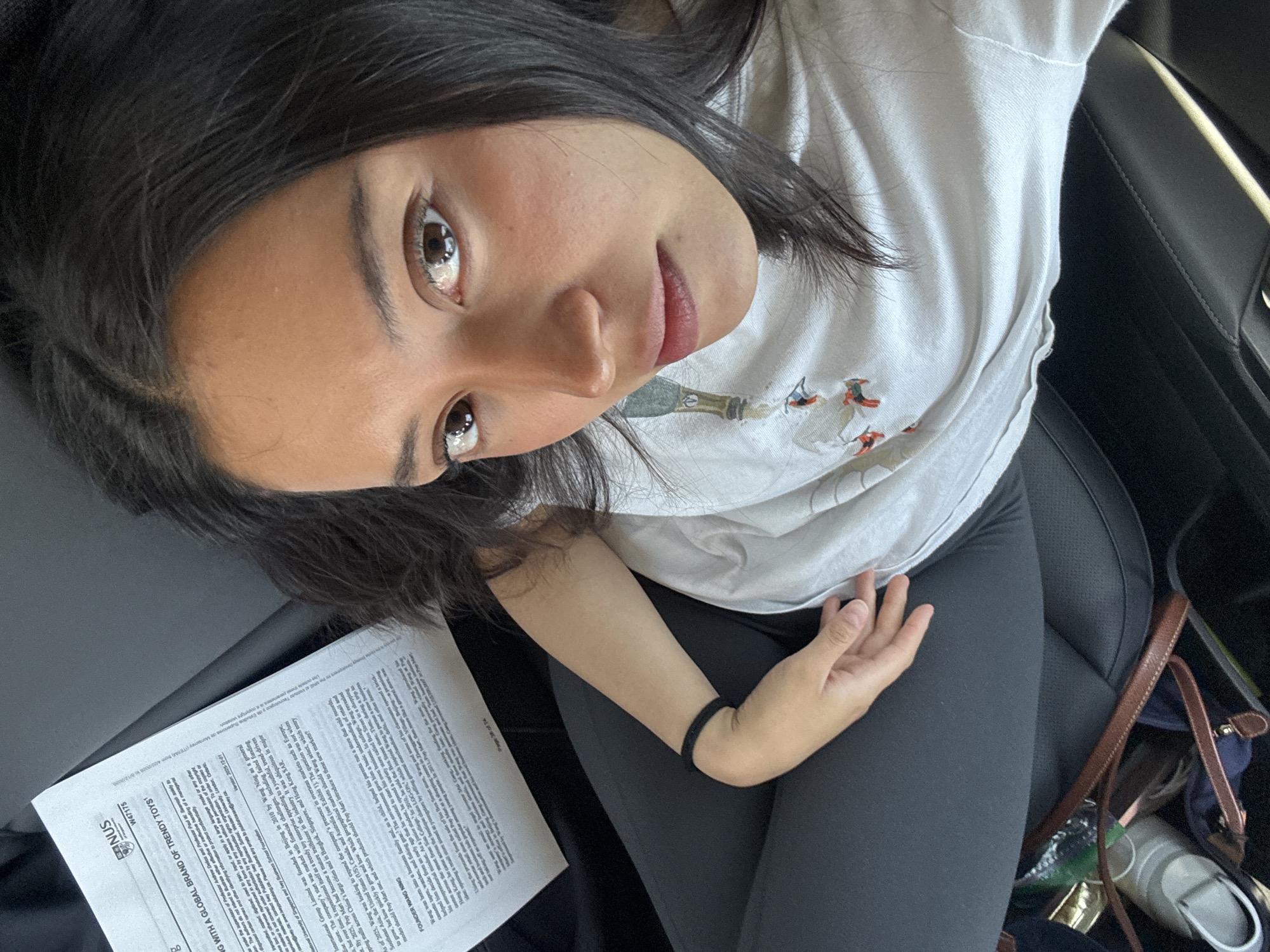 This media file captures a young woman seated in what appears to be the passenger seat of a car, likely in Puebla, Mexico, based on the provided context. She is the central subject, with dark hair, dark eyes, and a neutral to slightly soft expression, looking directly up towards the camera. She wears a casual white t-shirt featuring a subtle graphic design and dark leggings. A black band or hair tie is visible on her left wrist.
The setting suggests she is inside a vehicle, indicated by the black, possibly leather, car seat upholstery visible behind her and to her right. To her left, on the seat next to her, rests a white document, appearing to be a multi-page printed text or case study. Below her right arm, part of a brown leather bag strap, keys, and possibly a bottle or container are visible, suggesting personal belongings.
The primary activity is the woman posing for or taking a photograph, indicated by her direct gaze at the camera from an overhead angle. The lighting suggests it is daytime.
Visible text on the document includes "NUS", "W47175", "NG WITH A GLOBAL BRAND OF TRENDY TOYS¹", "FOUNDER WANG NING", "Pop Mart", and "Version: 2005-12-01". The text discusses the founding of "Pop Mart" in Beijing in 2010 by Wang Ning, its expansion into various global markets including Singapore, Hong Kong SAR, Europe, and the US, and mentions "University of Singapore". Towards the bottom right, partially visible text refers to "Tecnologico y de Estudios Superiores", further supporting the potential Mexican context for the image. The document also indicates "Page 3 of 14".