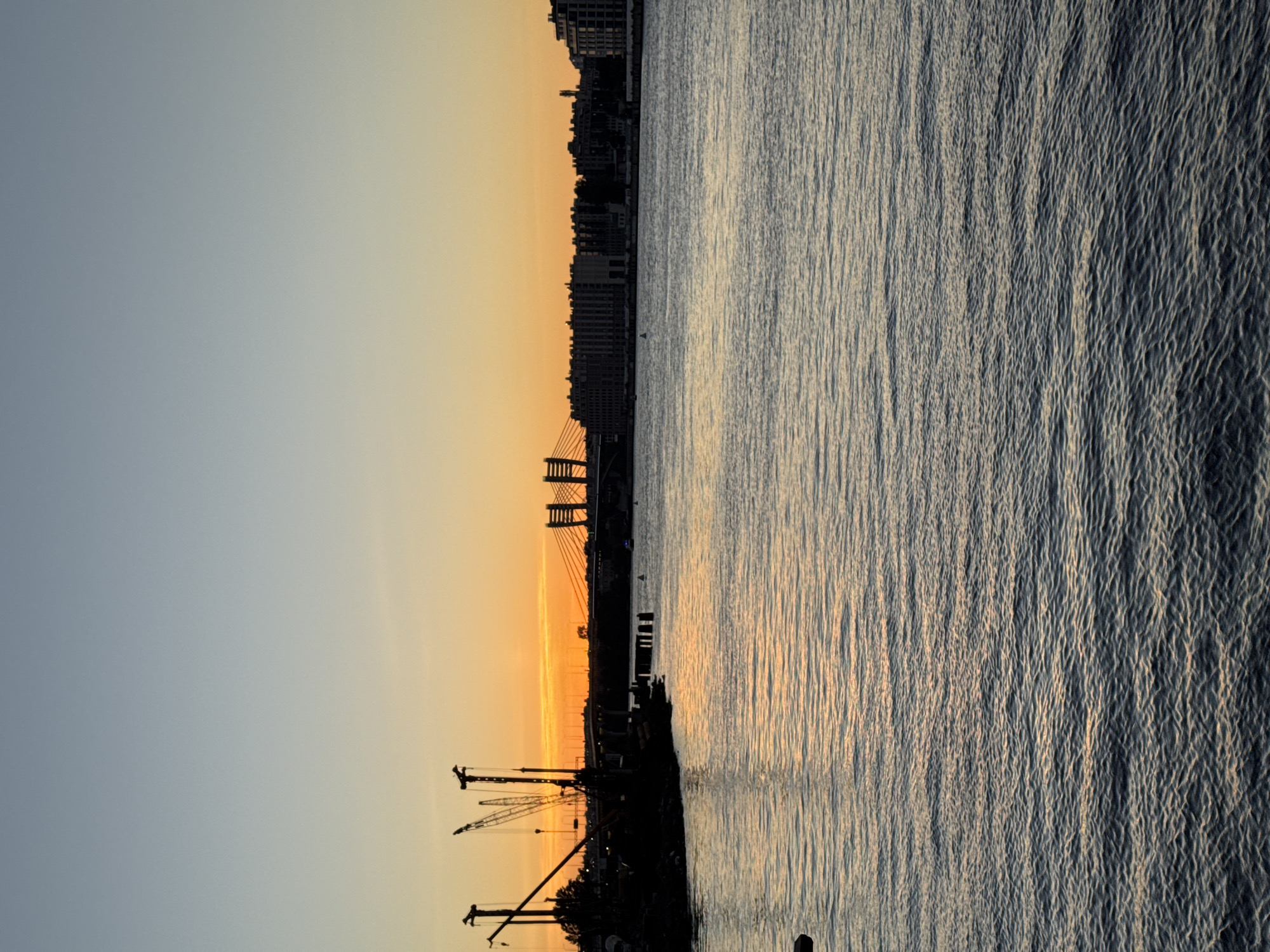The image captures a tranquil scene on a body of water in Saint Petersburg, Russia, during sunset. In the foreground, the water's surface is gently rippled, reflecting the warm hues of the sky. On the left bank, silhouetted against the vibrant orange and yellow sunset, are industrial elements, including cranes and what appears to be a dock or construction area. Further in the distance, a modern cable-stayed bridge spans the water, connecting the city's infrastructure. The far bank is lined with a cityscape of buildings, also silhouetted against the fading light. The sky transitions from a pale blue at the top to a fiery orange and yellow near the horizon, indicating the end of the day. The overall atmosphere is serene and industrial, highlighting the juxtaposition of nature and urban development in Saint Petersburg.