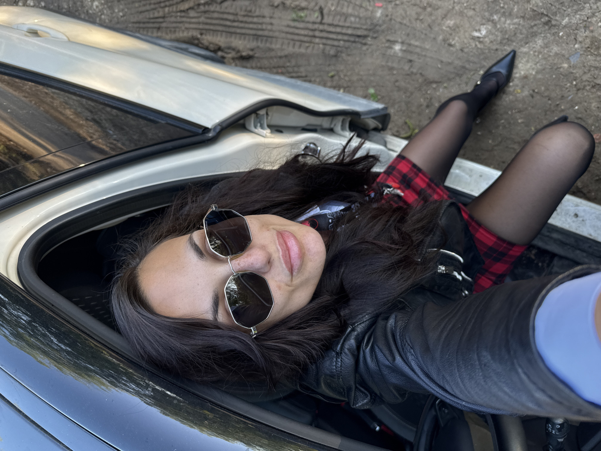 A young woman with long, dark hair and sunglasses is lying inside the trunk of a car. She is wearing a black leather jacket, a red plaid skirt, and black stockings with high-heeled shoes. The car appears to be parked on a dirt surface, possibly near a road or in an open area. The exact location in the city of Pleven, Bulgaria is not discernible from the image.