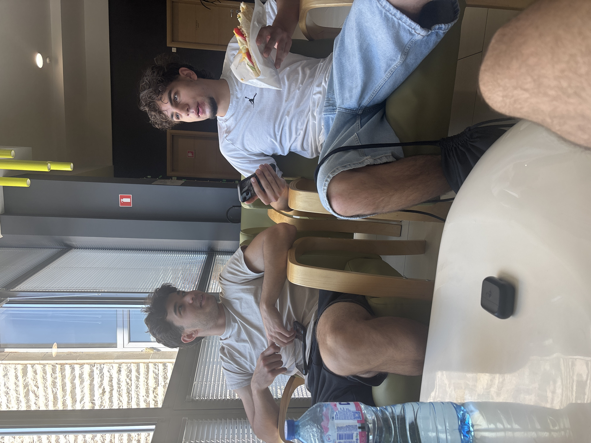 The image depicts two young men seated indoors, likely in a cafe or lounge area in Blagoevgrad, Bulgaria, during the daytime. The room is brightly lit by natural light streaming through large windows.
The man in the foreground on the right side of the frame (when oriented correctly) has dark curly hair and a goatee. He is wearing a white t-shirt with a black Jordan logo and light-wash denim shorts. He is seated in a green-cushioned chair with a wooden frame, holding a sandwich wrapped in white paper in his left hand and a smartphone in his right. He is looking directly at the camera.
The second man, positioned further back and to the left, also has dark curly hair and is wearing a light-colored t-shirt and dark shorts. He is seated in a similar green-cushioned chair and is looking towards the left, out of the window.
In the immediate foreground, a part of a person's leg and thigh is visible, along with a white table surface. On this table are a plastic water bottle with a blue cap and a small black rectangular object, possibly an earbud case.
The background features modern interior elements: bright fluorescent lights on the ceiling, yellow cylindrical decorations on a wall, and a dark grey wall with a red fire extinguisher sign. Behind the first man, there are dark wooden panels or cabinets. The large windows, fitted with horizontal blinds, offer a view of a sunny exterior with a light-colored, textured stone building facade and a clear blue sky. The overall scene suggests a casual and relaxed break. The only clearly visible text is the Jordan logo on the first man's t-shirt.