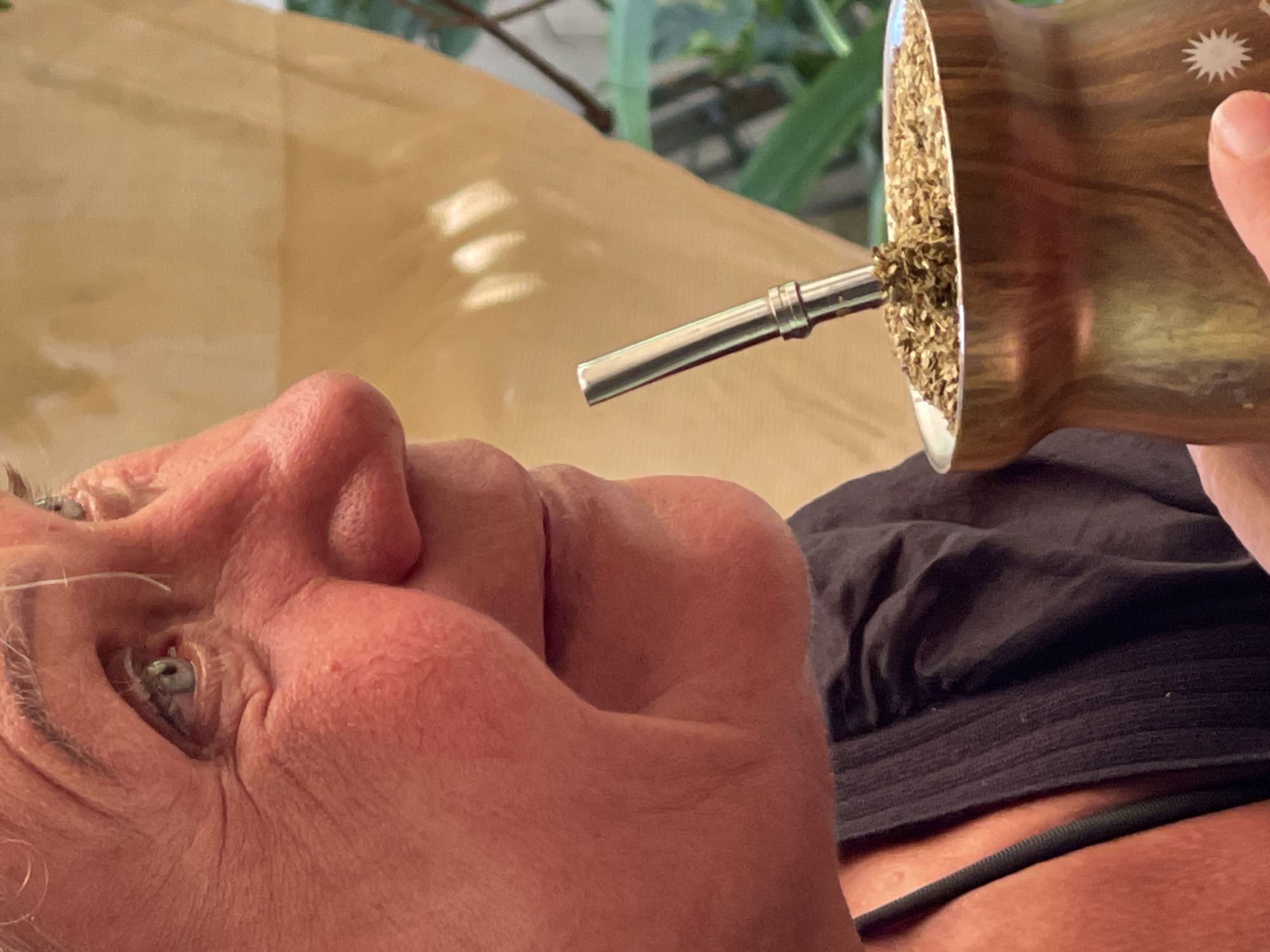 An extreme close-up shot shows the profile of an older woman's face, with her light blue eyes looking to the left. She has fair skin with visible wrinkles and rosy cheeks. Her mouth is slightly parted as she sips from a mate gourd. The gourd, made of a metallic material, is filled with dried yerba mate leaves and has a silver straw, called a bombilla, inserted into it. The woman is wearing a dark top. The background is blurred, showing a warm, yellowish surface and hints of green foliage, suggesting an outdoor or well-lit indoor setting. The image does not provide any information about the specific location in Đà Nẵng, Vietnam.