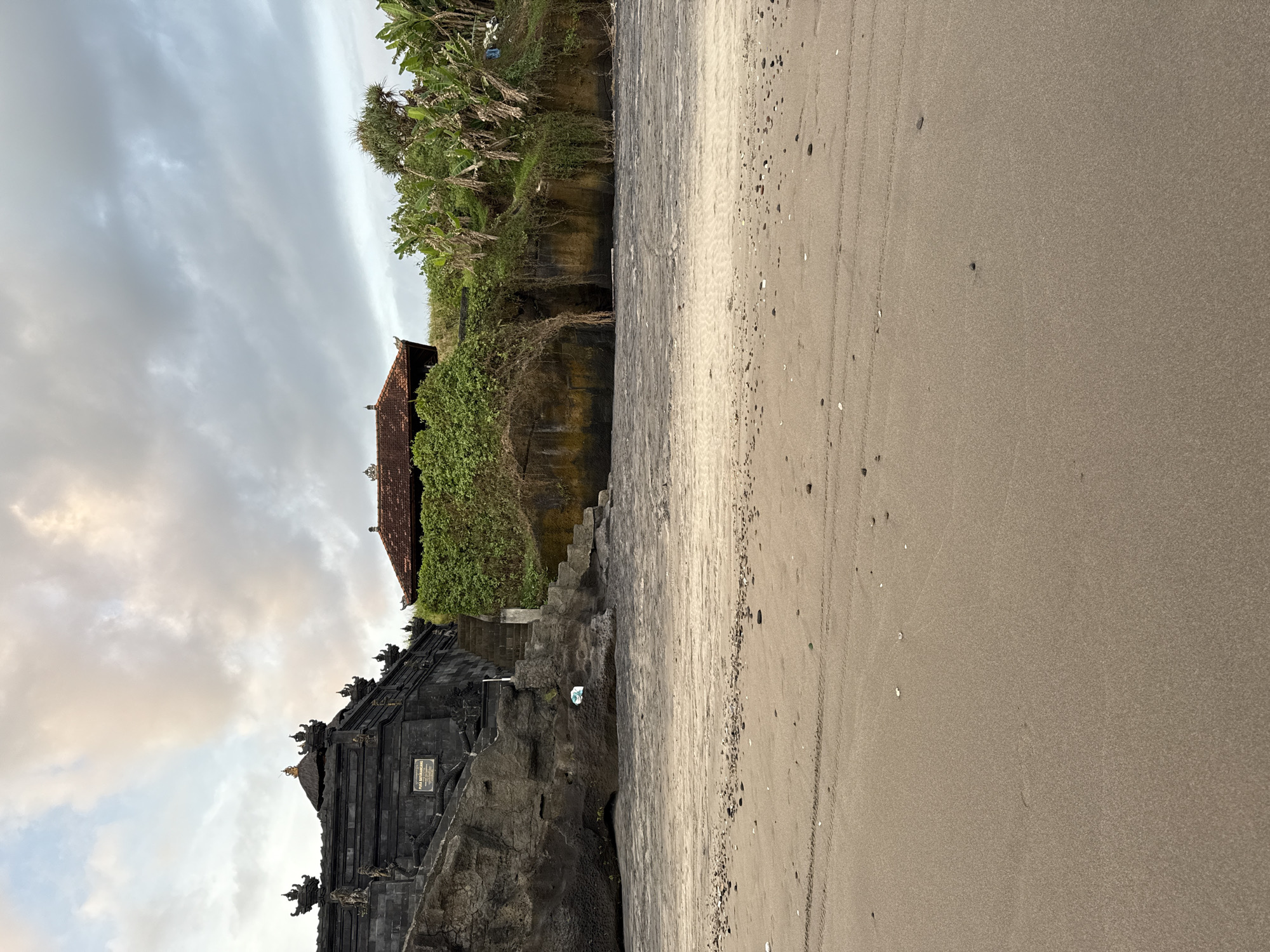 The image depicts a beach scene with a Balinese temple on a rocky cliff. The temple is dark in color and has traditional architectural details. Adjacent to the temple, a structure with a red tile roof is visible. Dense green vegetation covers the top of the cliff. The beach is sandy and extends towards the right side of the image. The sky is overcast with a mix of gray and white clouds. The location is Unknown, Unknown.