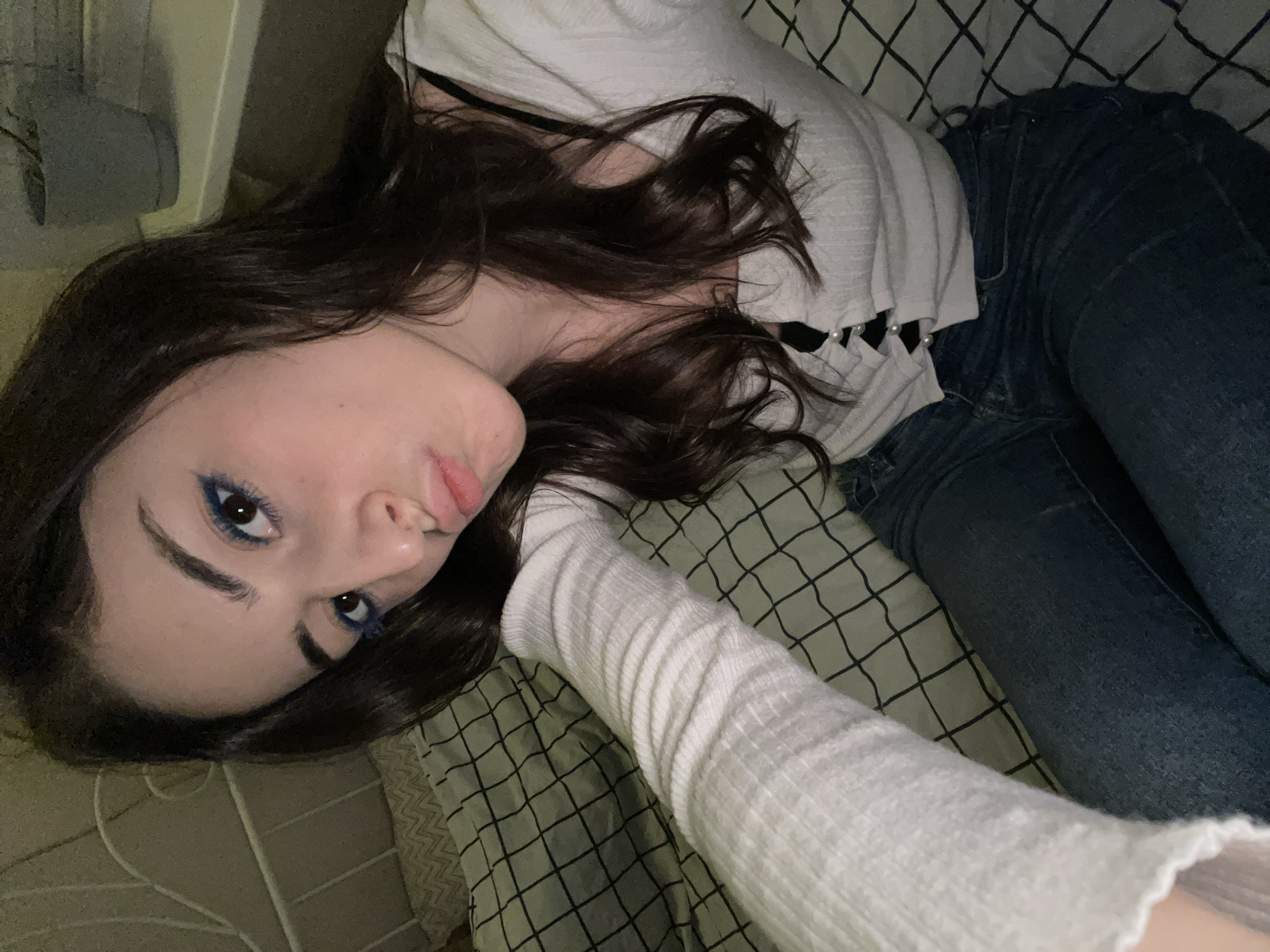 The image shows a young woman lying on a bed with a patterned blanket, possibly in a residential setting. She has dark hair and is wearing blue eyeliner. She is wearing a white top with pearl details and blue jeans, her right arm is extended wearing a white, long-sleeved shirt. The lighting is dim. There is no information in the picture to suggest this is in Moscow, Russia.