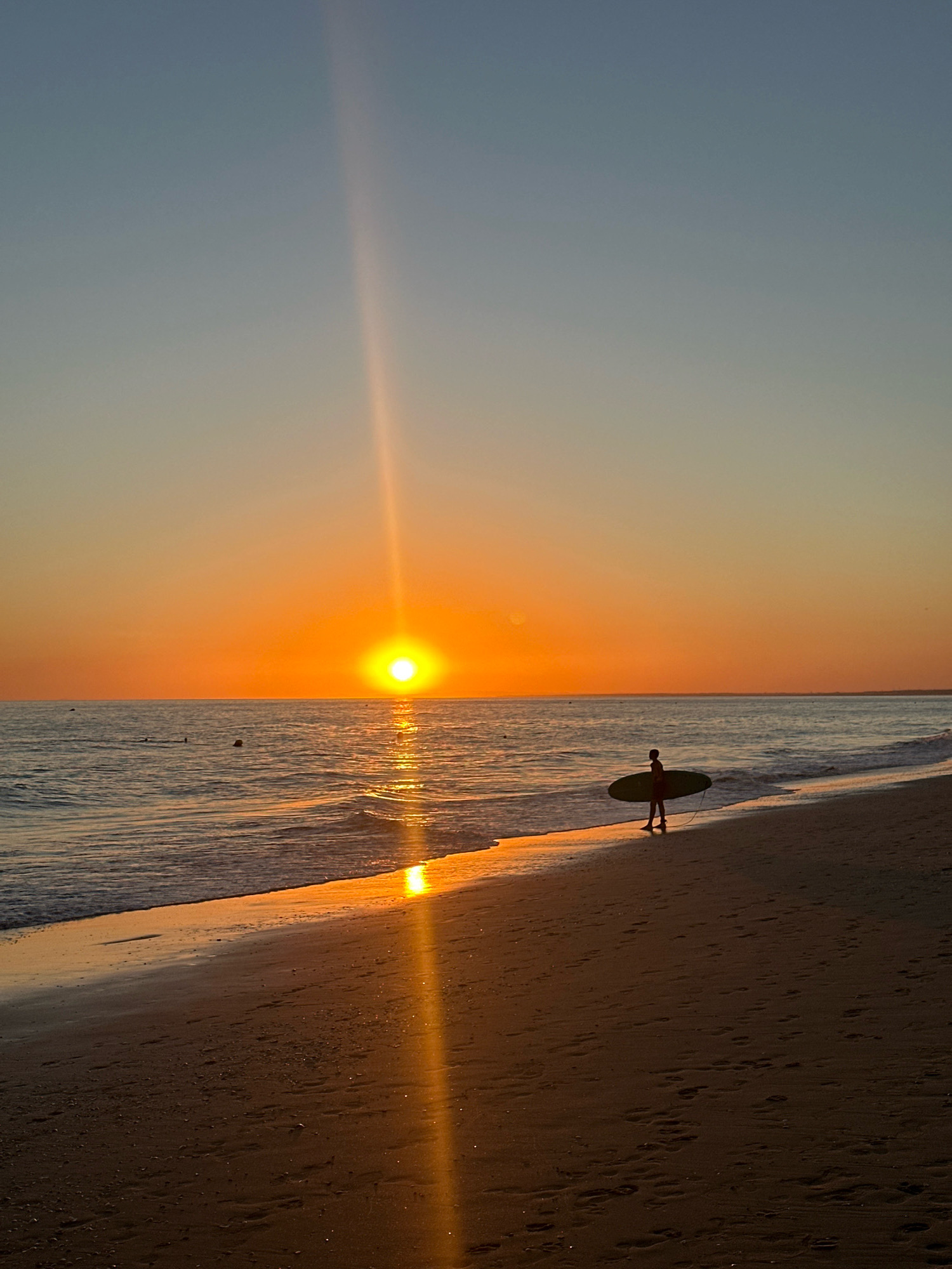 This image depicts a beach scene in Punta Umbría, Spain, during sunset. The sun is low on the horizon, casting a warm, orange glow across the sky and reflecting on the rippling ocean waves. A single person, silhouetted against the light, walks along the wet sand near the water's edge, carrying a surfboard. The sand is textured with footprints. In the distance, a few other individuals are visible in the water. The sky transitions from a pale blue at the top to a vibrant orange near the horizon. A distinct beam of sunlight pierces the sky vertically from the sun.