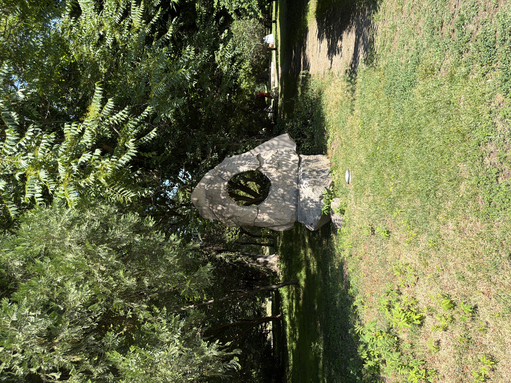 A medium shot captures a stone sculpture with a circular opening in the center, situated in a sun-dappled park. The sculpture is in the foreground, on the left side of the frame. It is light gray and appears to be made of rough, weathered stone. Behind the sculpture, a dirt path winds to the right, leading into a wooded area. Tall trees with dense green foliage form a canopy overhead, casting shadows on the grass and path. Sunlight filters through the leaves, creating a dappled lighting effect. The grass is a mixture of green and dry, yellowed patches. In the background, beyond the path, a few people are visible, appearing as small figures enjoying the park. One figure on the right is wearing a red top, and another on the far right has a light-colored garment, possibly a tent or umbrella. The overall impression is a serene park scene on a sunny day, with the unusual stone sculpture as a focal point. This scene is located in the City Park in Pazardzhik, Bulgaria.
