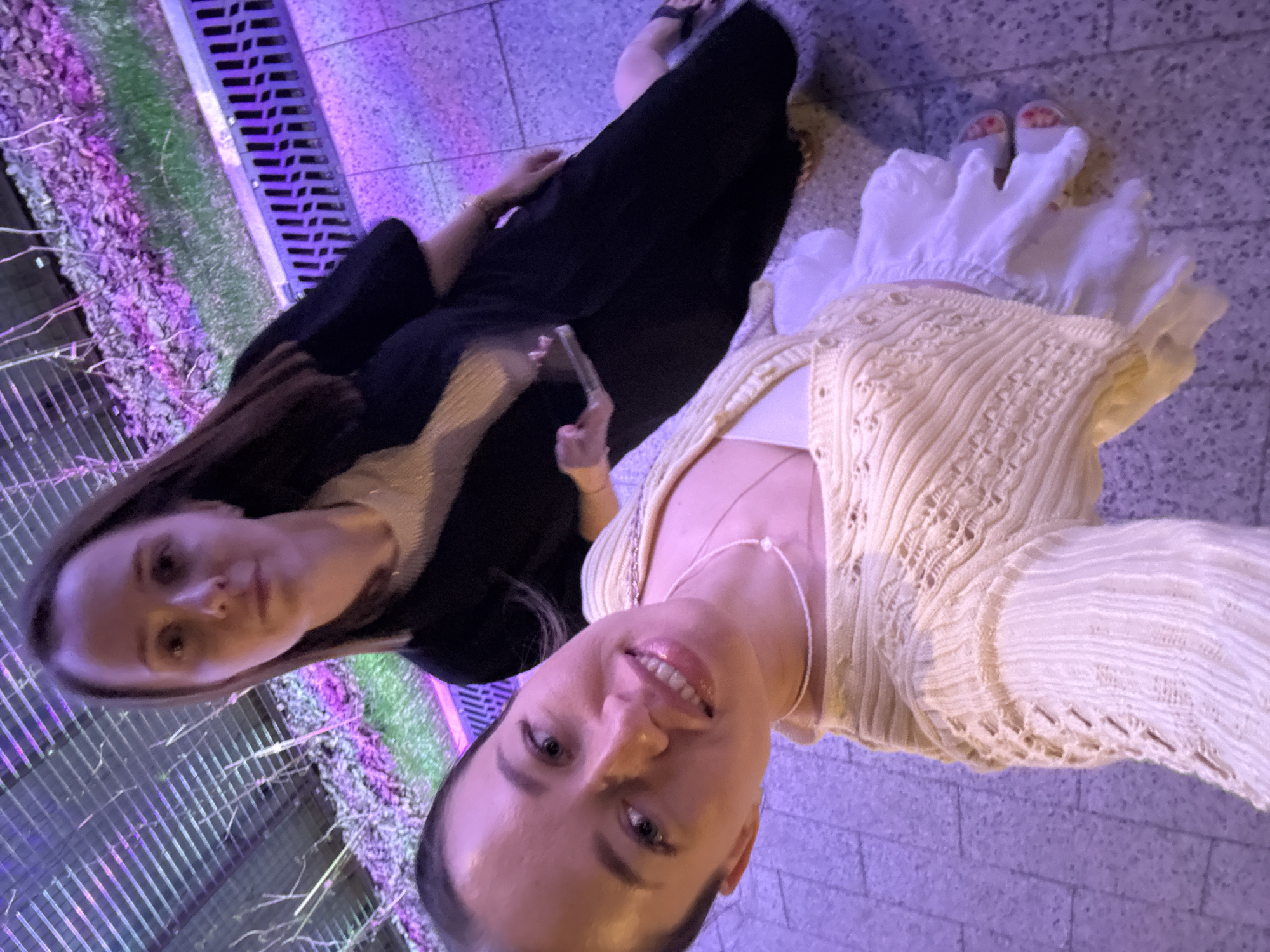 Two women are posing for a selfie in what appears to be an urban setting. The woman in the foreground is wearing a yellow sweater, a white top, and a pearl necklace. She is smiling at the camera. The other woman is wearing a black outfit. They are standing on a tiled surface. A grate with vegetation is on the left side of the image. The picture was taken in Perm, Russia.