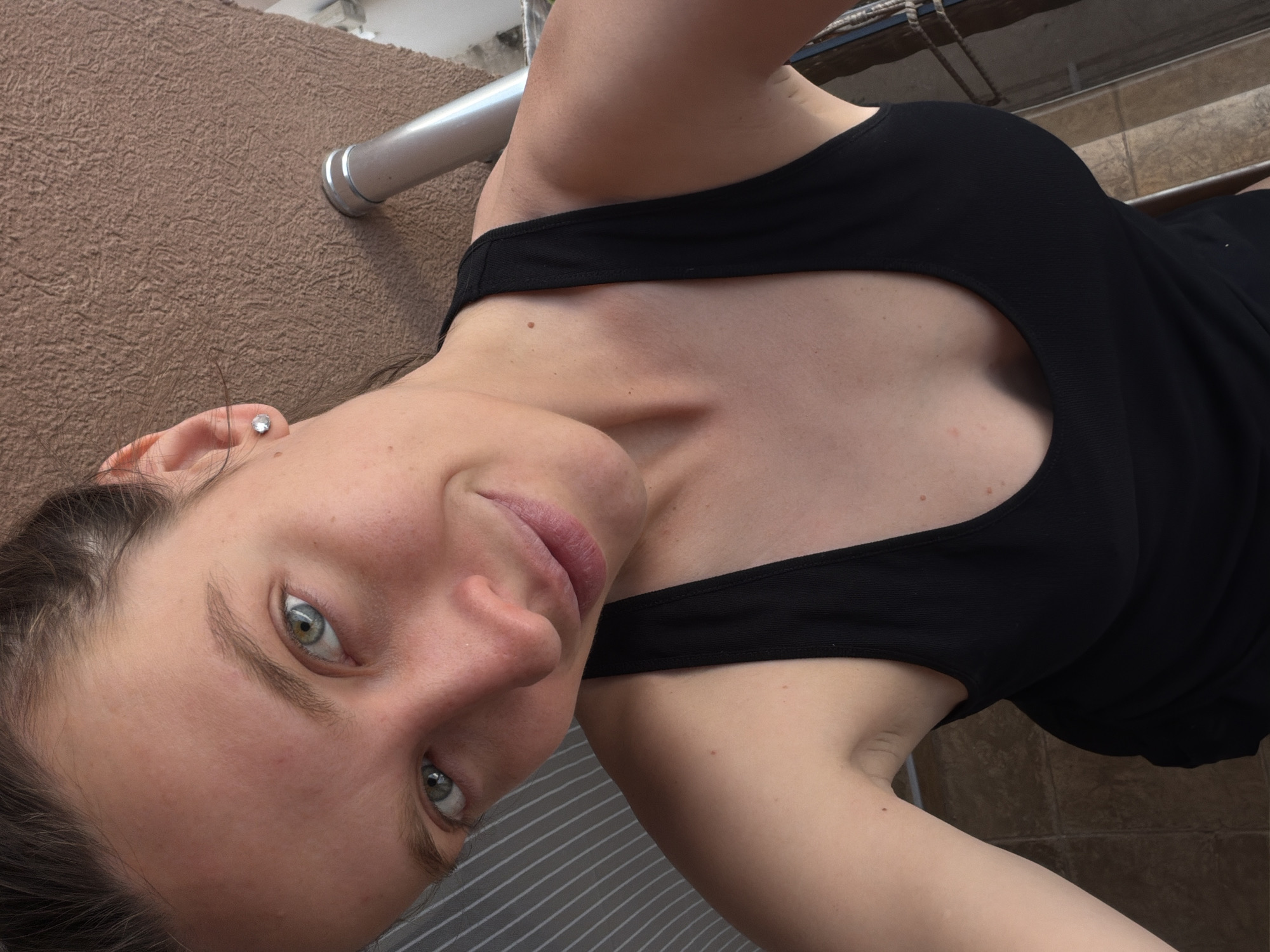 A woman with fair skin, blue eyes, and dark hair is captured from a low angle, looking directly at the camera with a slight smile. She is wearing a black tank top that reveals some of her chest and collarbone area. She has a small stud earring in her left ear.
The background is composed of different elements. To the left, there is a textured, brown wall. A silver metal pipe runs horizontally across the top-left quadrant of the frame. Beneath her, there's a grey surface with parallel lines or a striped pattern. Further back and to the right, the background transitions to a tiled floor with brown and beige tones.
There are no apparent details related to the city of Budva, Montenegro. There's nothing to suggest a specific outdoor location or any landmarks associated with Budva.