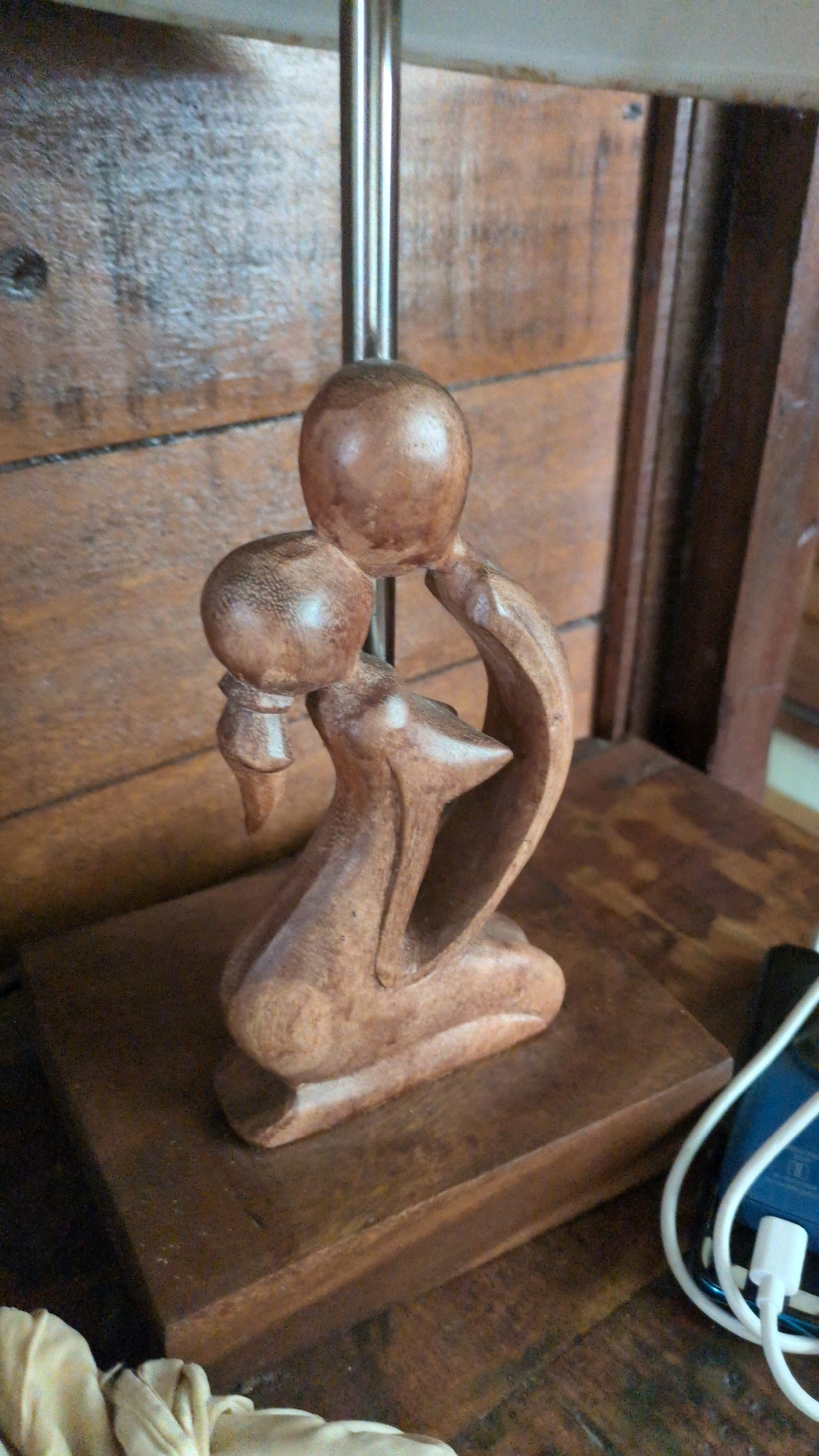 A wooden sculpture of two abstract figures embracing is positioned on a dark wooden base. The figures are carved in a flowing, organic style, with rounded heads and simplified bodies. The sculpture is lit from above, casting subtle shadows. Behind the sculpture, a wooden wall paneling provides a warm, textured backdrop. To the right of the sculpture, a blue smartphone with a white charging cable is visible, suggesting a contemporary setting. A light-colored fabric is draped in the lower left corner of the frame, adding a softer element to the scene. The overall impression is one of intimacy and artistry, with the natural materials contributing to a rustic yet elegant aesthetic. The specific location in Ubud District, Indonesia, is unknown from the image alone.