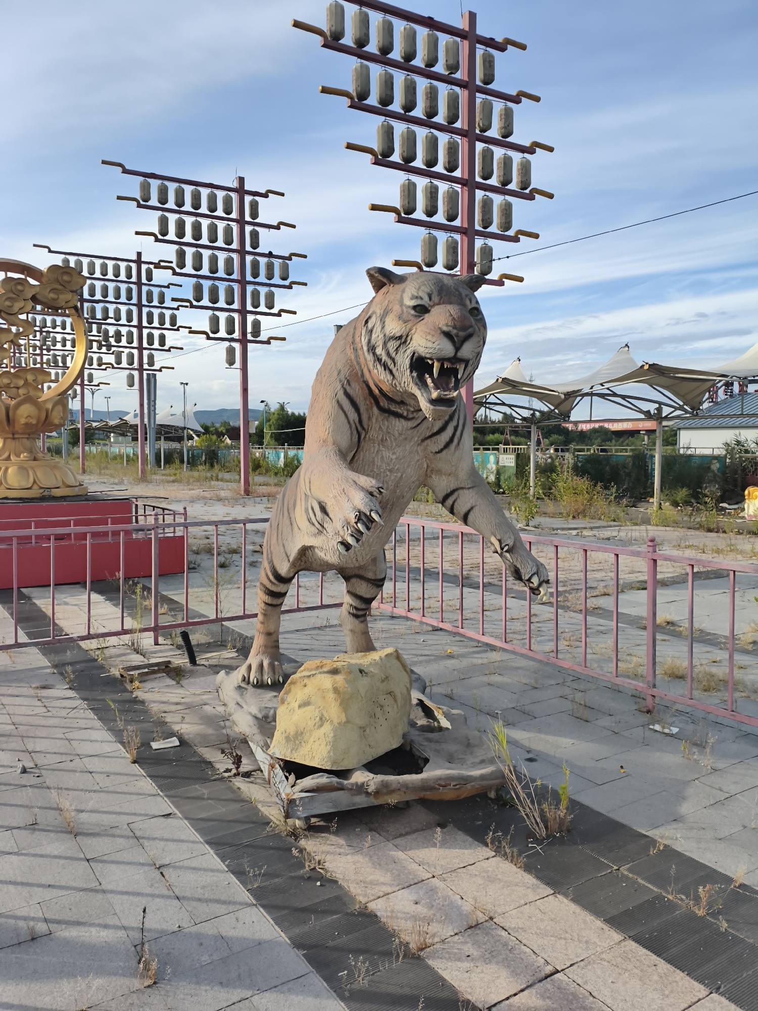 In the city of 新城村 (Xinchengcun) / 신성촌 (Sinseongchon), China, an outdoor scene features a large, weathered statue of a tiger in mid-pounce, positioned on a raised platform with a faux rock base. The tiger statue is light brown with black stripes and an open mouth revealing fangs. Behind the tiger, a golden, ornate structure with cloud motifs is partially visible on the left. In the background, several tall, red metal poles stand in rows, adorned with many small, cylindrical lanterns. The ground is paved with square tiles, some of which are cracked or uneven, with sparse weeds growing through the gaps. A pink metal fence encloses the area. Beyond the fence, a few white, tent-like structures are visible, along with trees and distant buildings under a partly cloudy blue sky. The overall impression is one of an abandoned or neglected outdoor attraction.